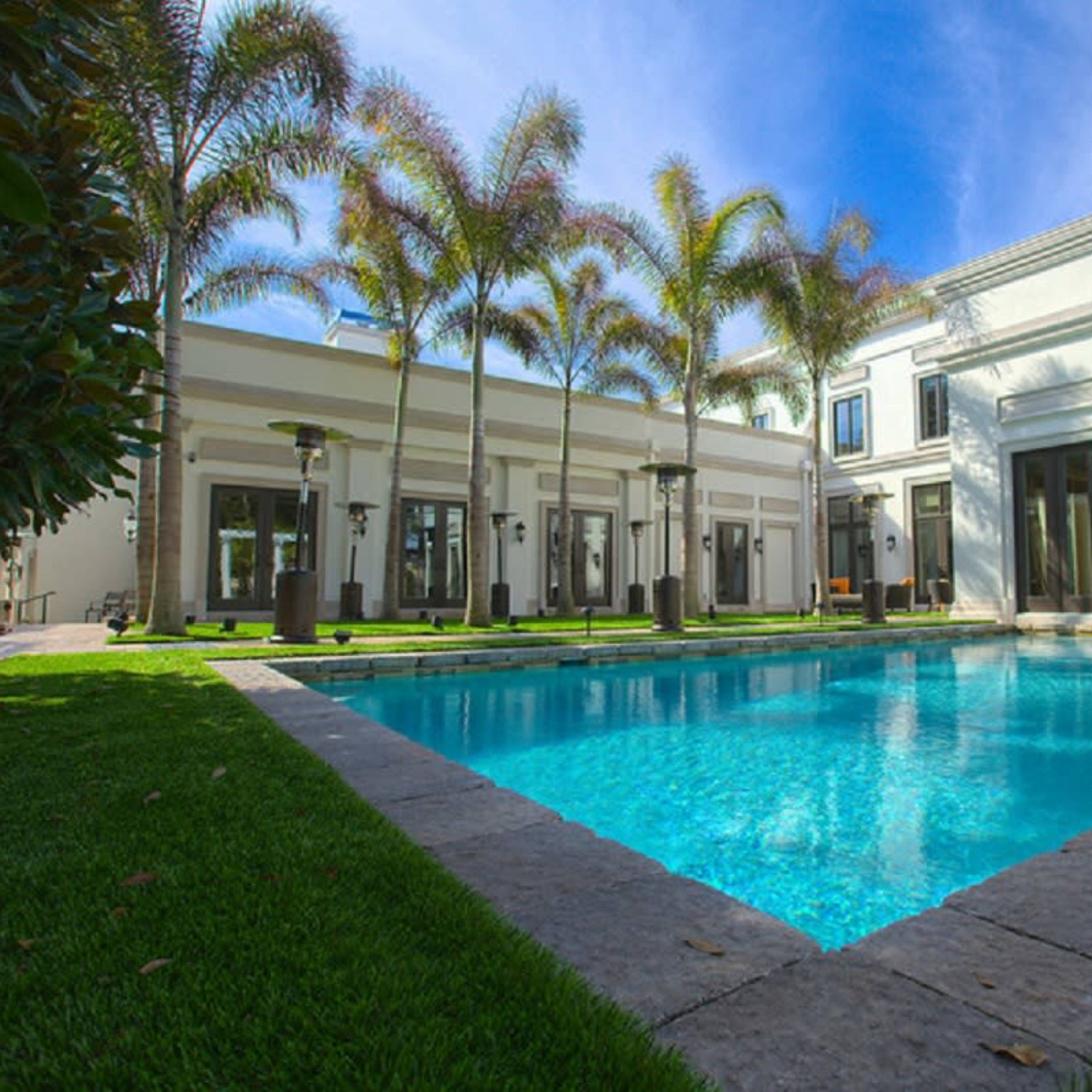 A clear blue swimming pool is surrounded by manicured grass and palm trees, with modern buildings in the background featuring large windows.
