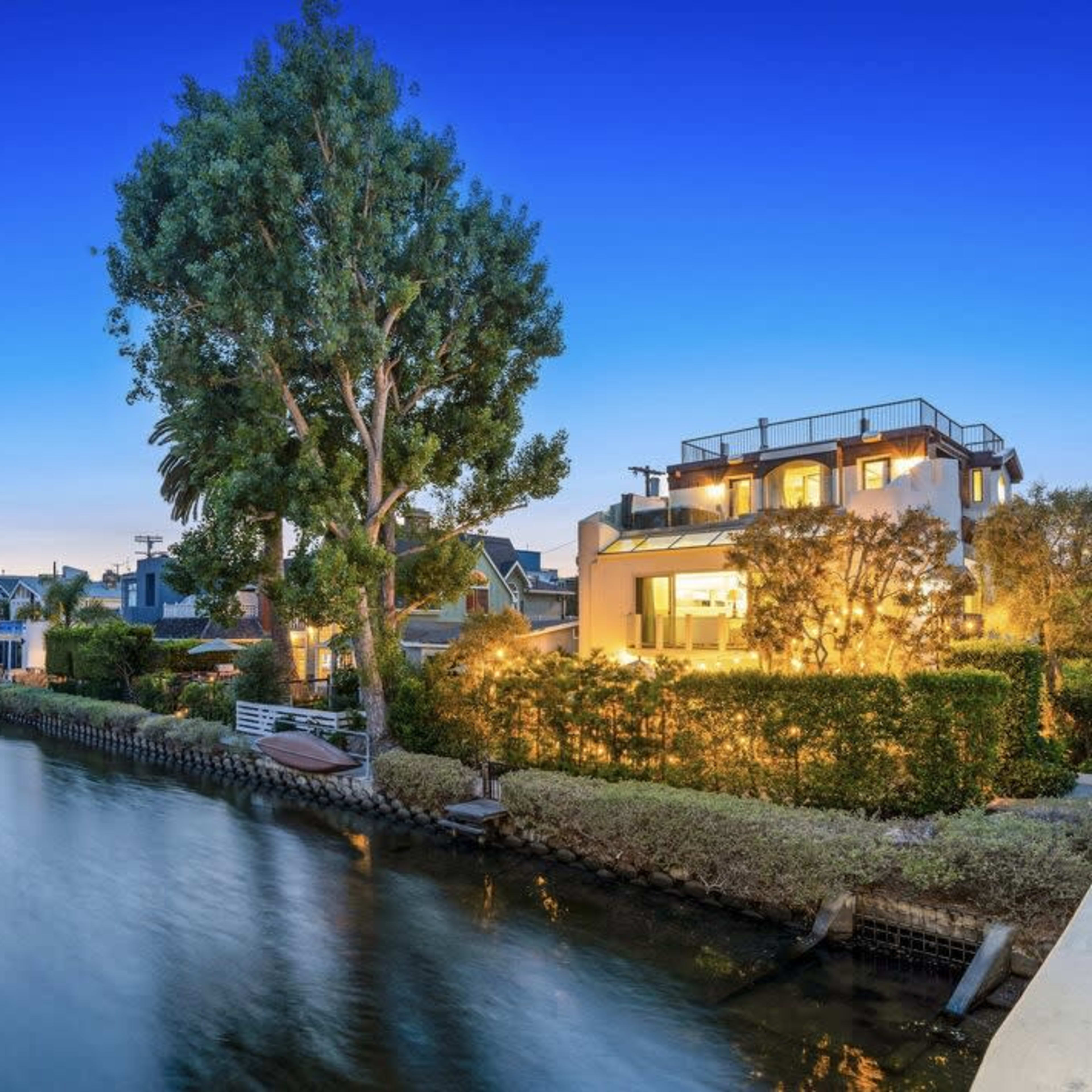 A serene canal lined with houses, illuminated in the evening twilight, with a large tree and boats visible along the water's edge.