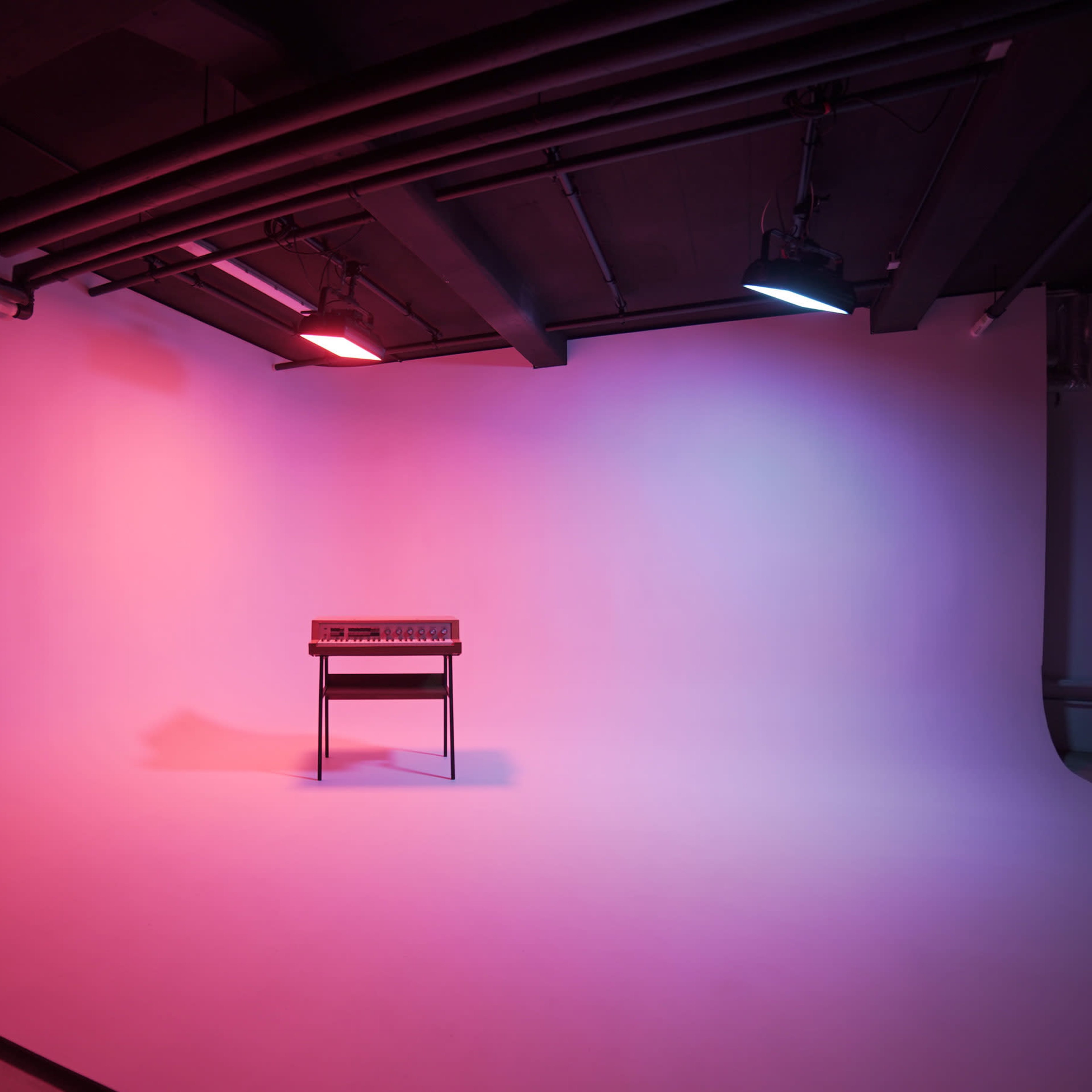 A keyboard is positioned on a stool in the center of an empty studio with colored lights illuminating the background.