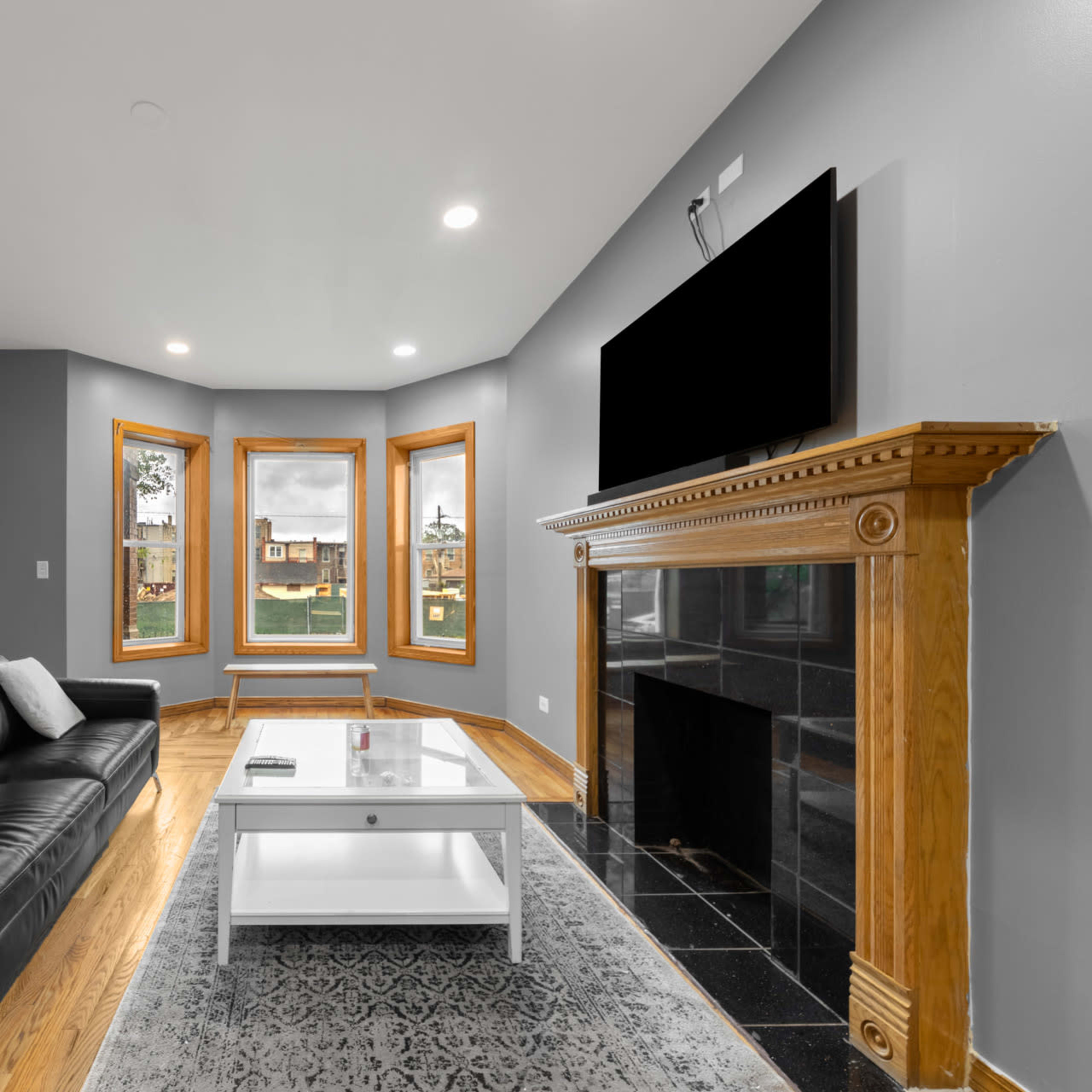 A living room features a black leather sofa, a glass coffee table, and a mantel with a large flat-screen TV mounted above a fireplace, surrounded by three windows showcasing an outdoor view.