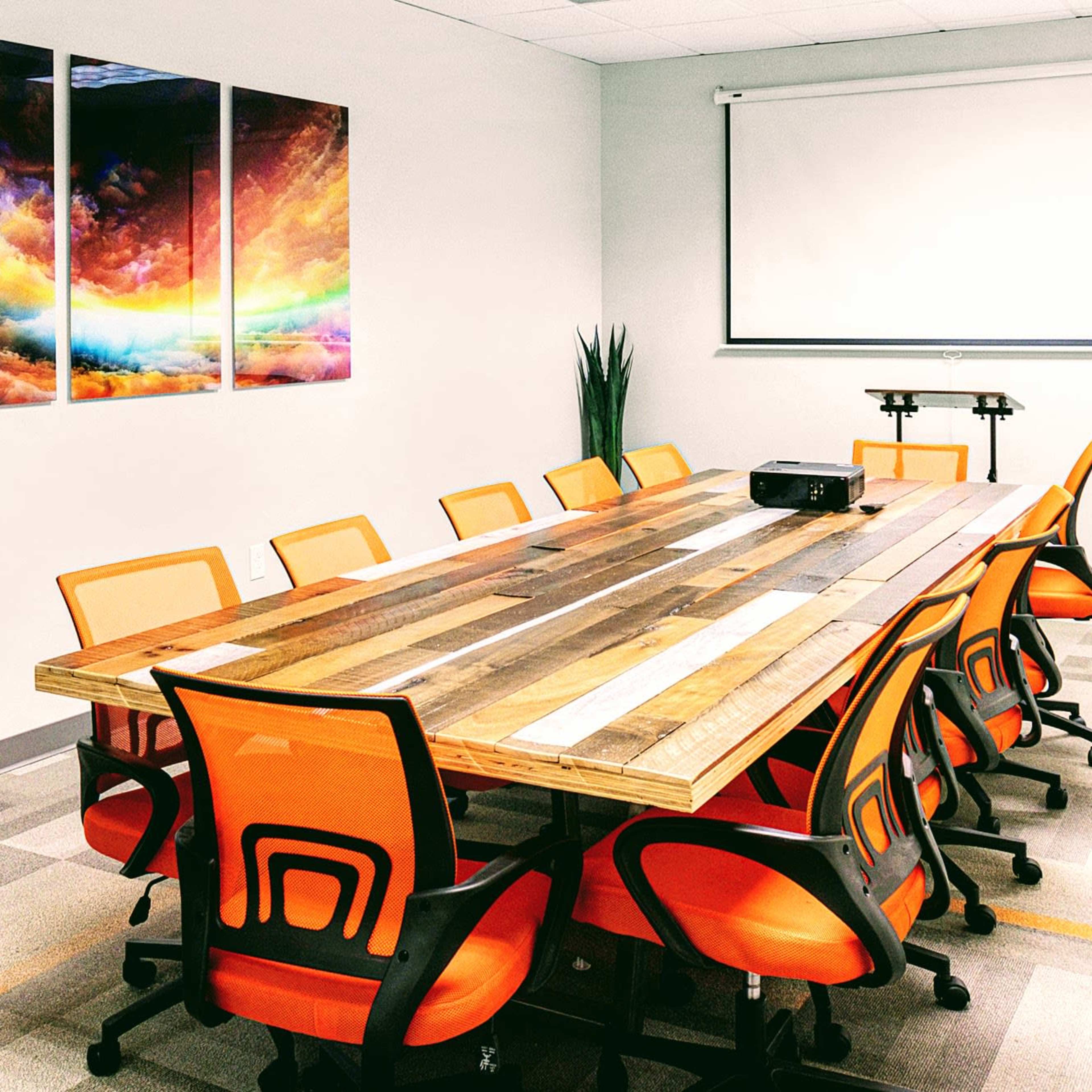A conference room with a large wooden table surrounded by orange chairs, featuring a projector and a colorful abstract artwork on the wall.