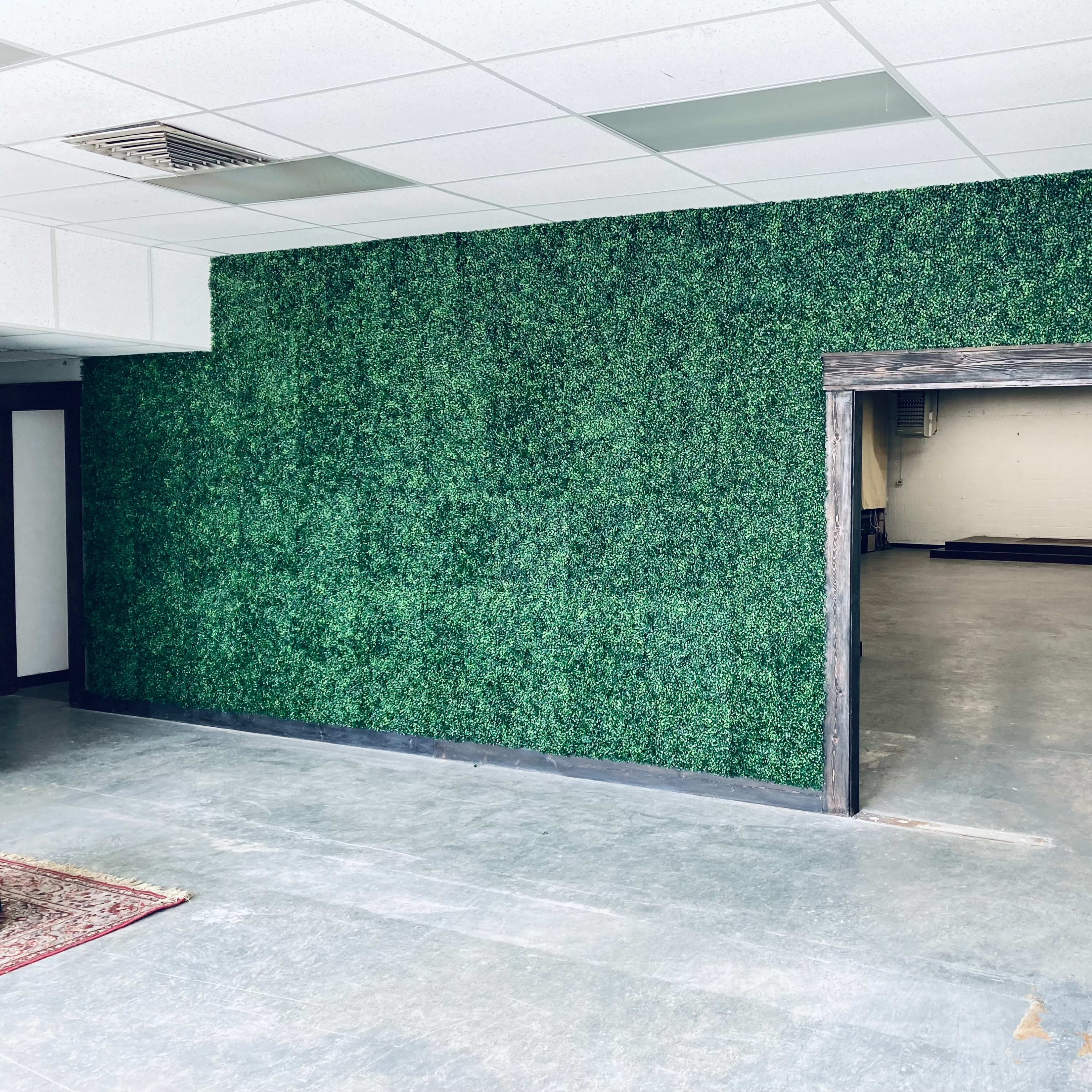 A room with a green artificial hedge wall and an open doorway leading to a space with a concrete floor and minimal furnishings.
