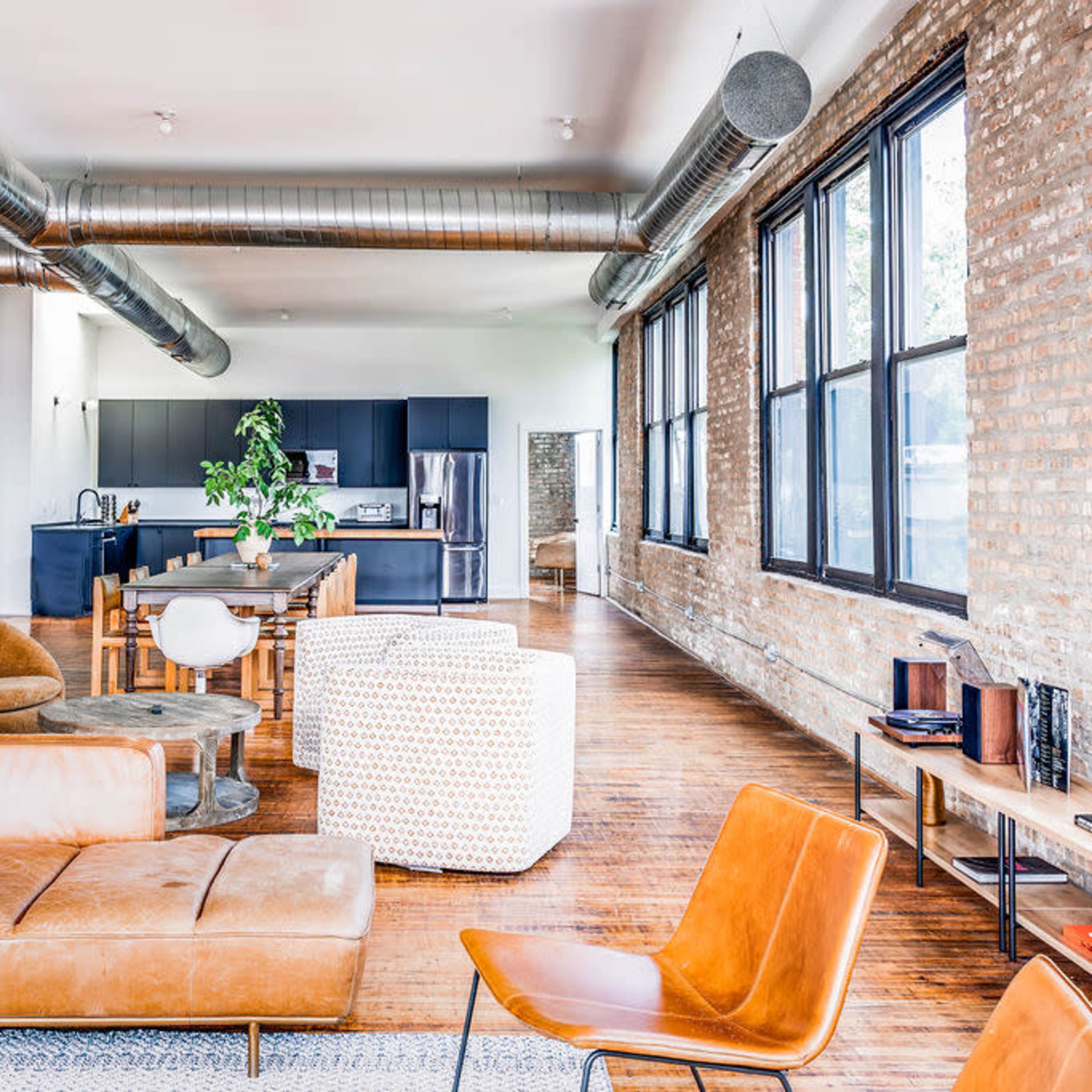A spacious, modern living area featuring large windows, brick walls, and various seating options including a leather sofa and chairs.