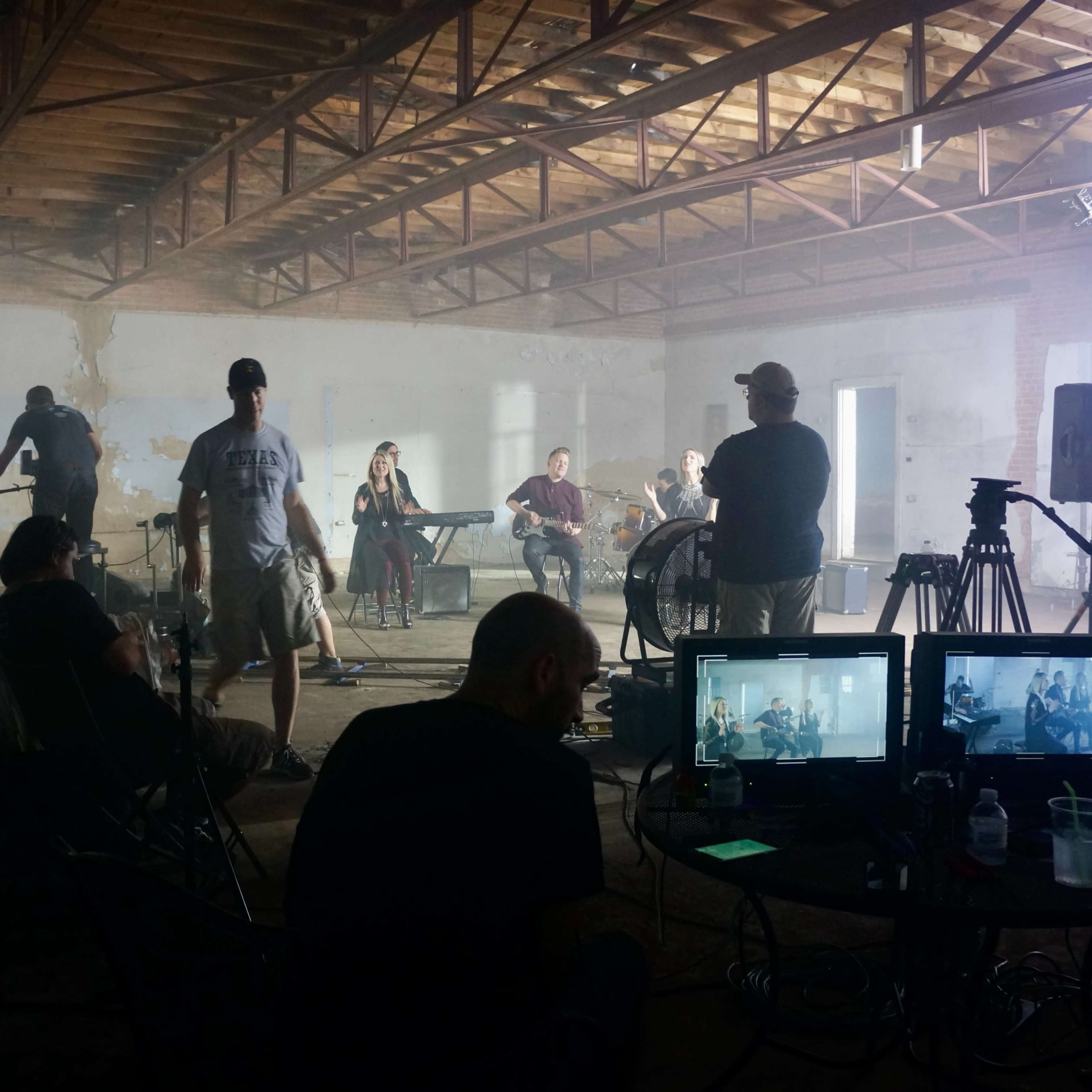 A group of musicians performs in a dimly lit warehouse while cameras and equipment are set up for recording.
