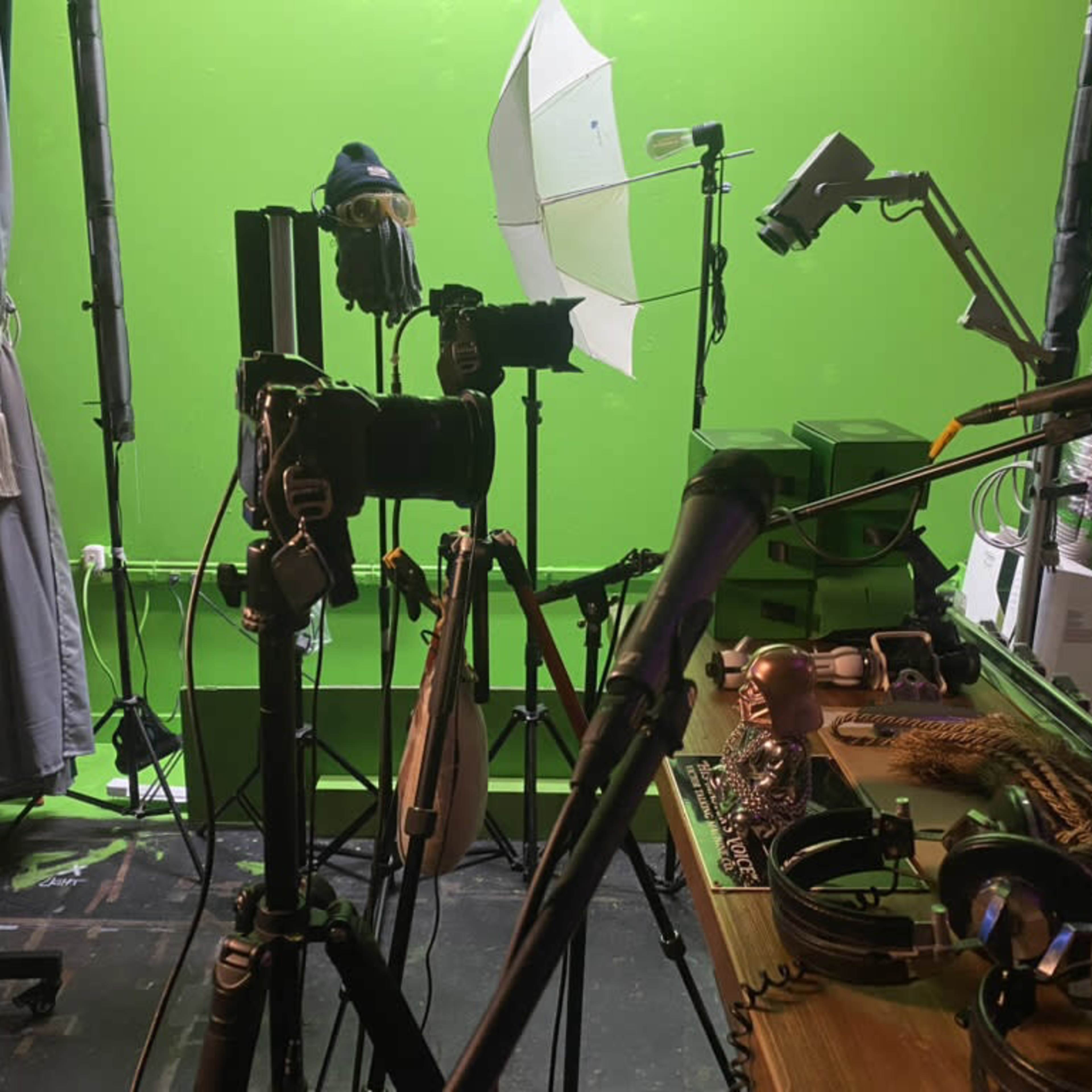 A green screen studio is set up with cameras, microphones, and lighting equipment placed on tripods near a wooden table displaying various accessories.