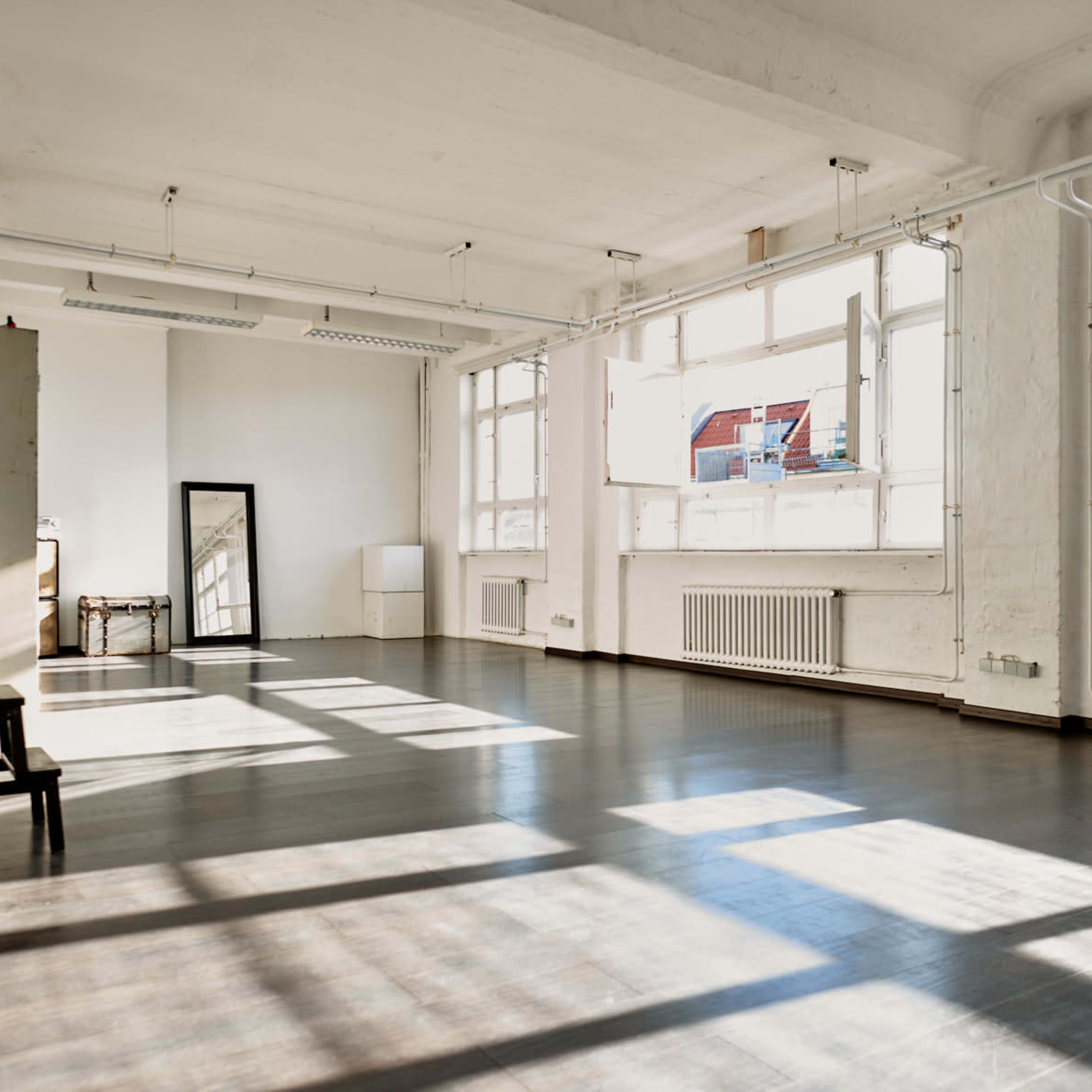 A spacious, well-lit interior room with large windows and hardwood flooring, featuring a mirror and a few pieces of furniture.