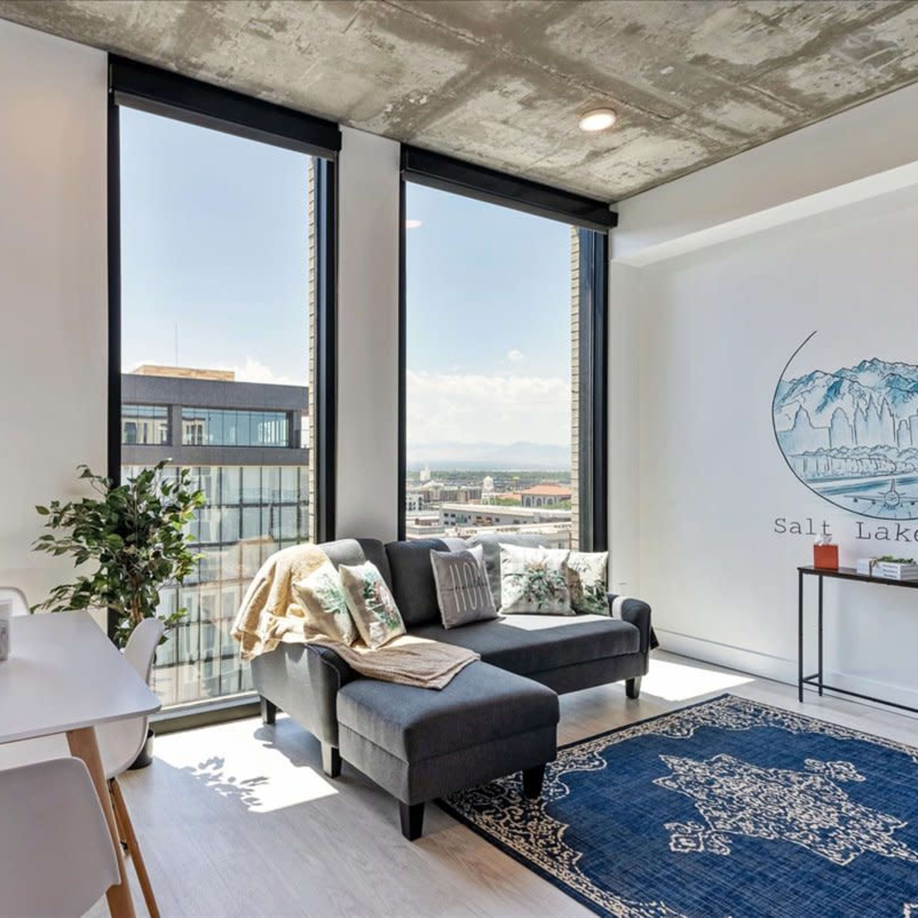 A modern living space with large windows, a gray sofa, a dining table, and a decorative wall featuring a Salt Lake City mural.