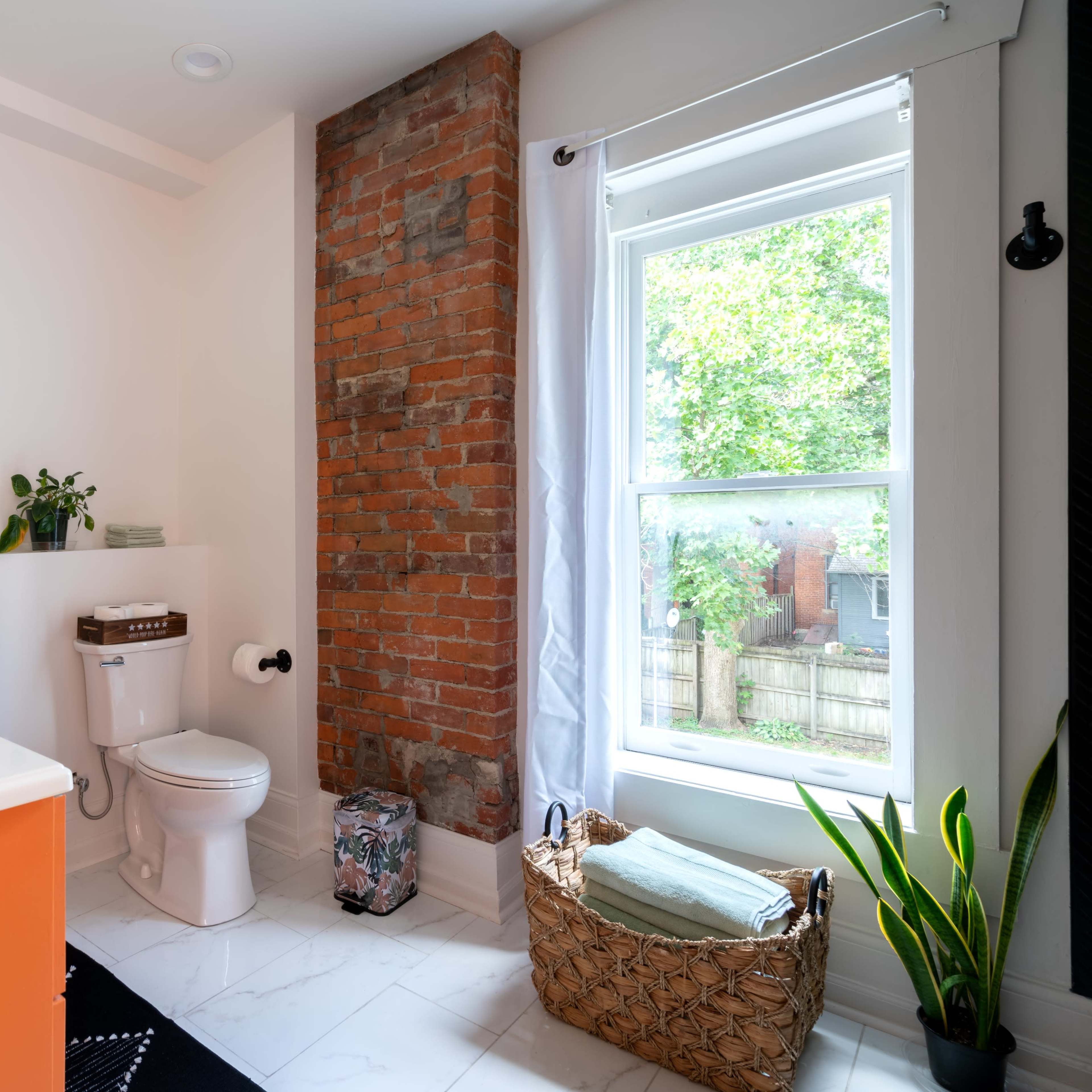 A modern bathroom features a combination of exposed brick and sleek fixtures, with a window overlooking a garden and a decorative basket in the corner.