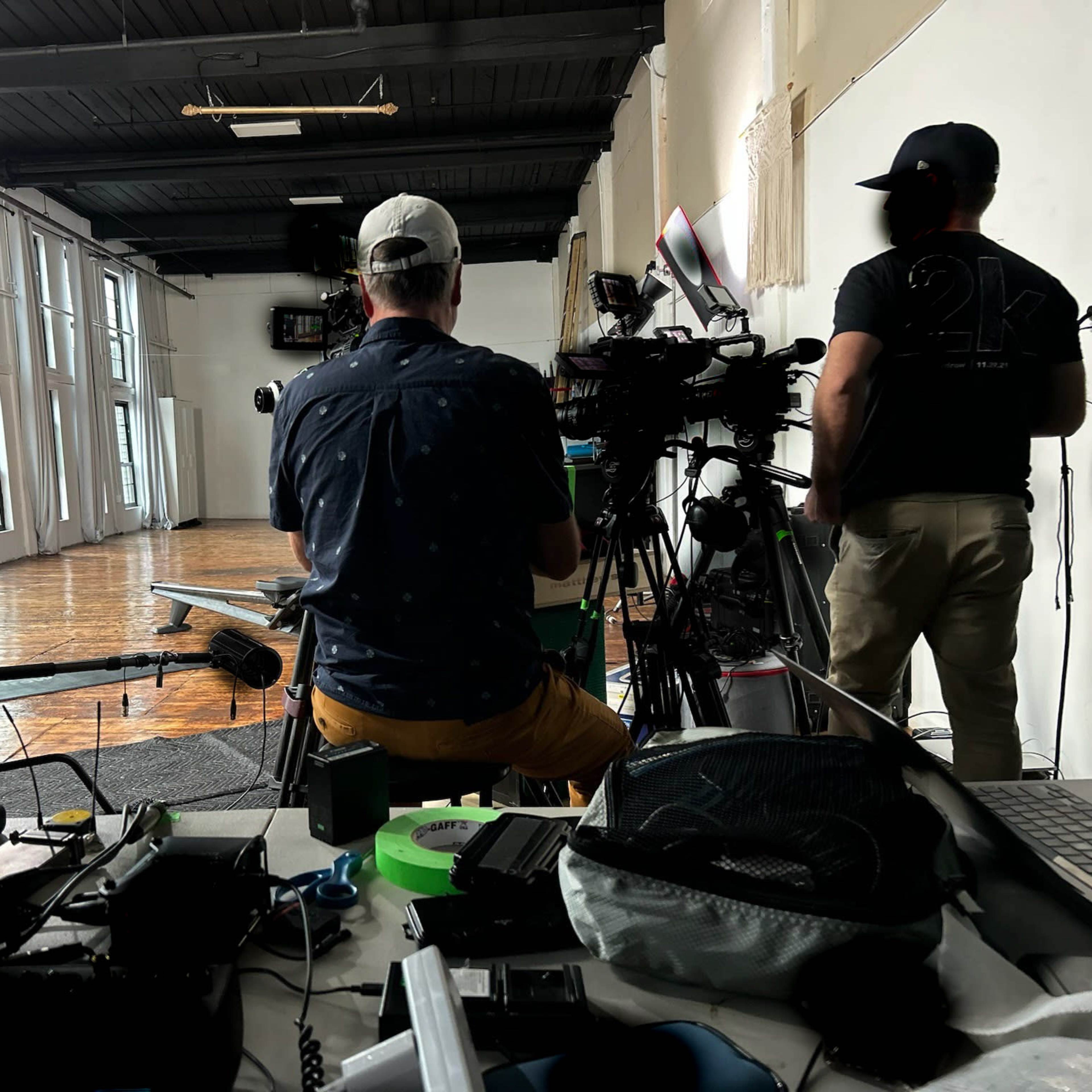 A filming setup in a spacious room with large windows, featuring a camera crew, equipment on a table, and a person on the floor preparing for a shoot.