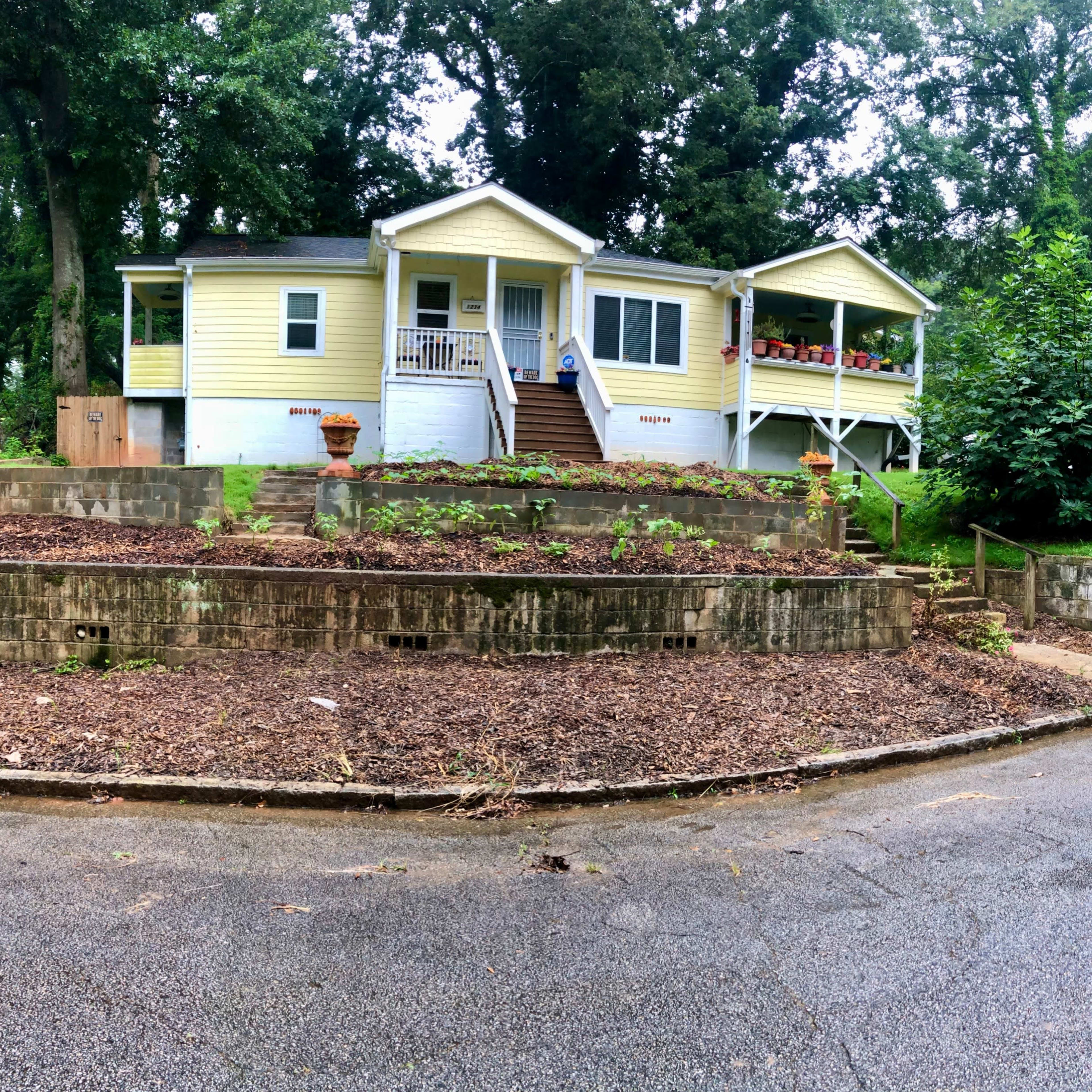 A yellow house with a front porch, situated on a landscaped yard with mulch and a gravel pathway, alongside a curved road.