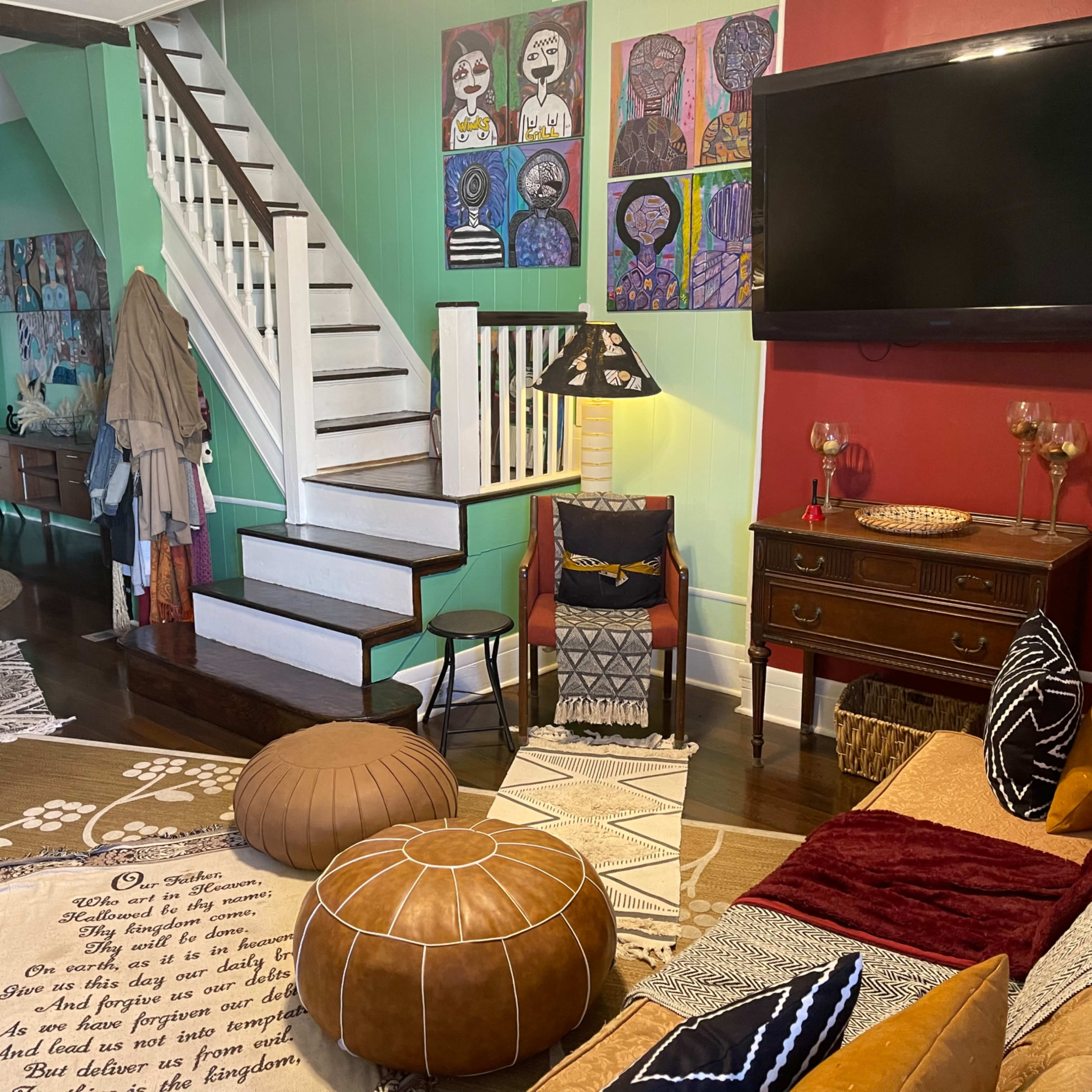 A cozy living area with a staircase, various decorative artworks on the walls, a television, a couch with colorful cushions, and a patterned area rug.