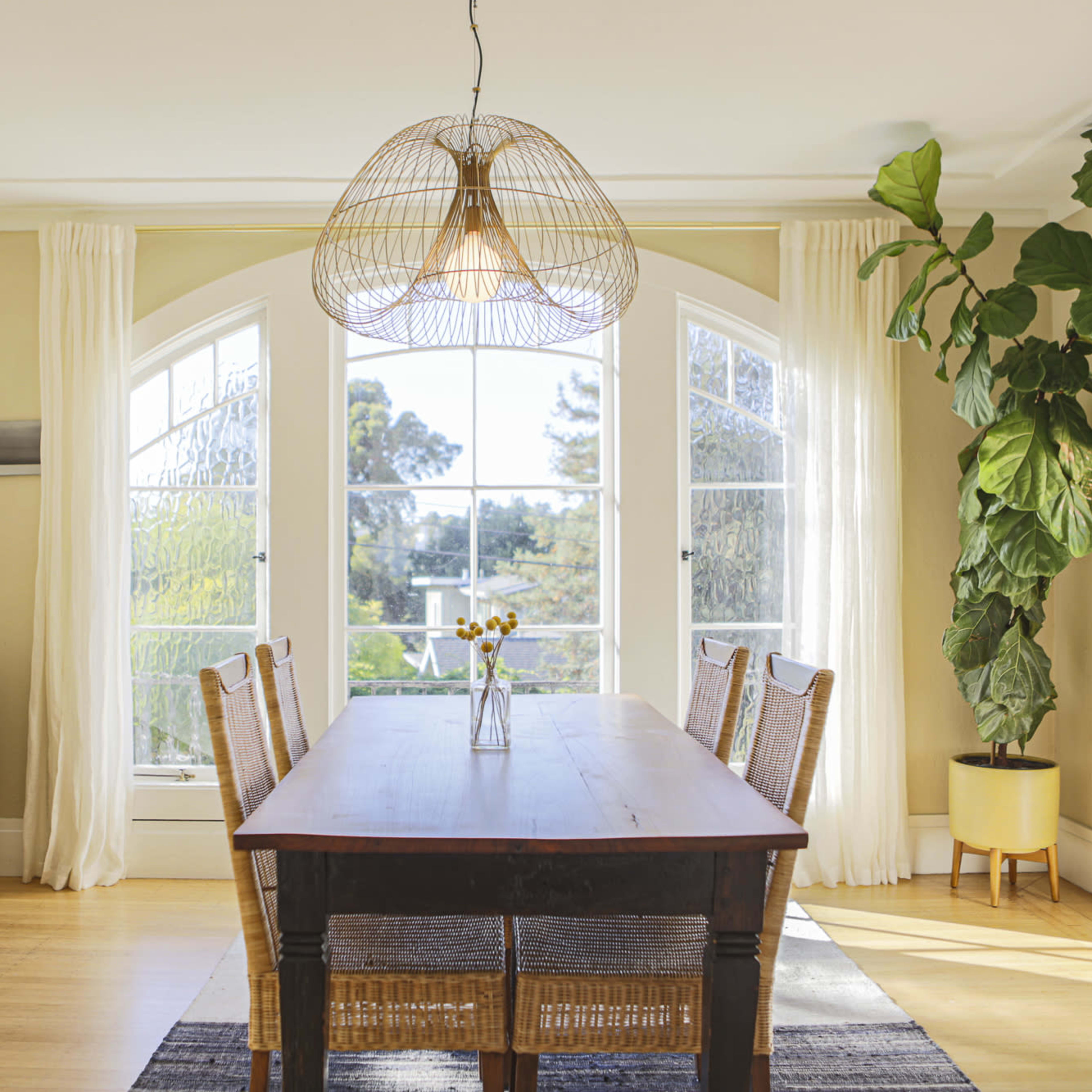 A wooden dining table is centered in a light-filled room with large windows and a plant in the corner.
