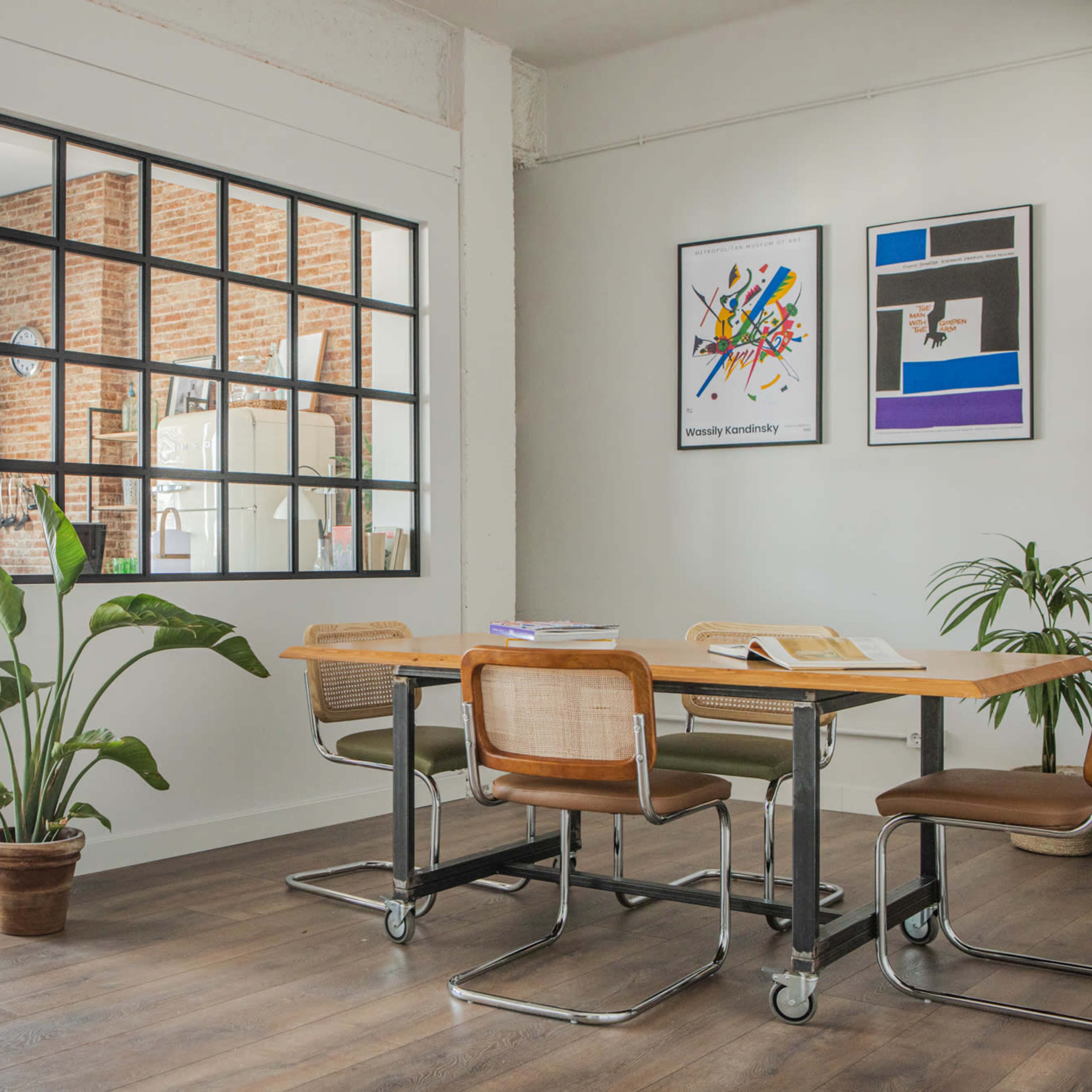 A modern interior featuring a dining area with a wooden table and chairs, separated from a kitchen by a glass partition, with plants in the foreground.