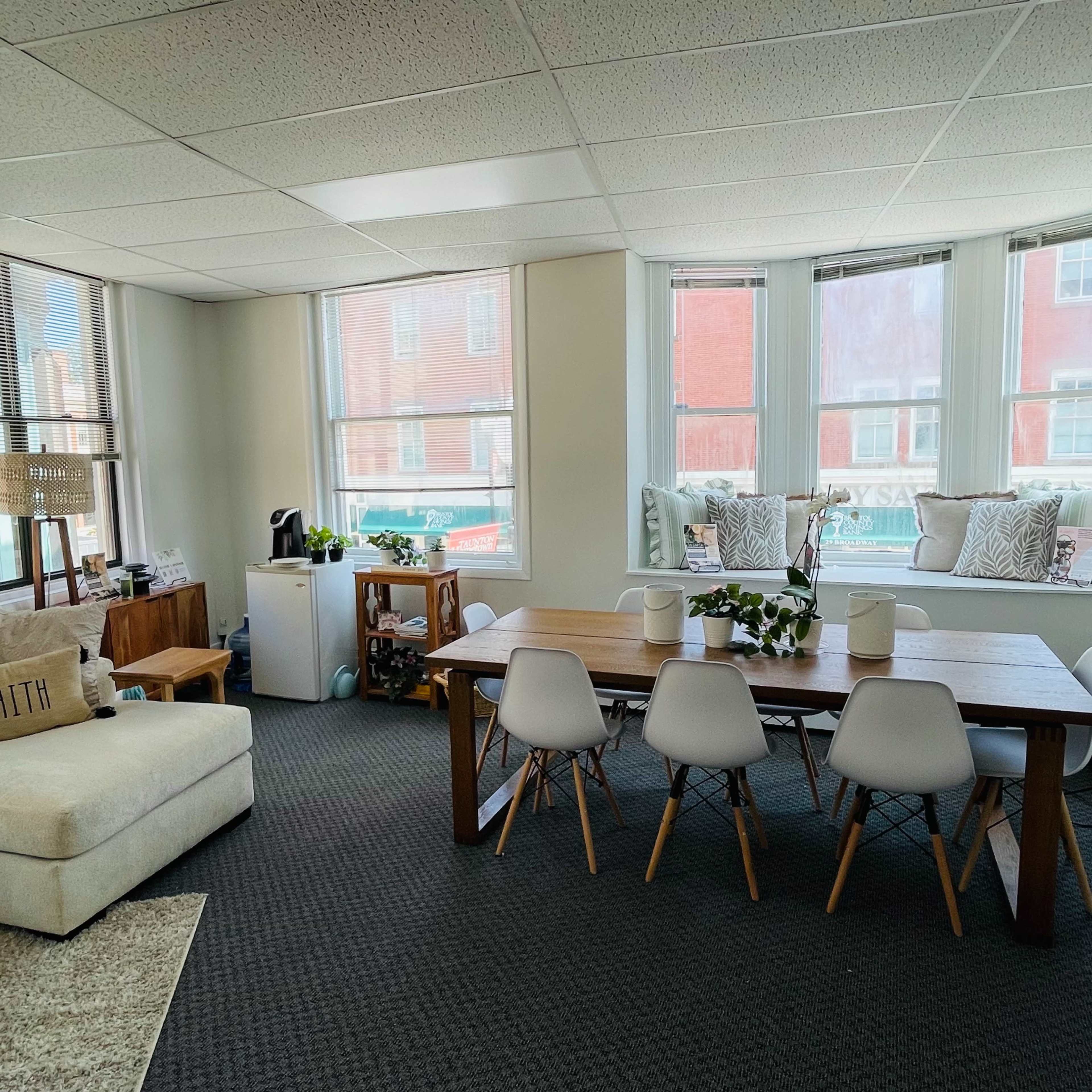 A modern living area features a light-colored sectional sofa, a wooden dining table with white chairs, and large windows with cushions.
