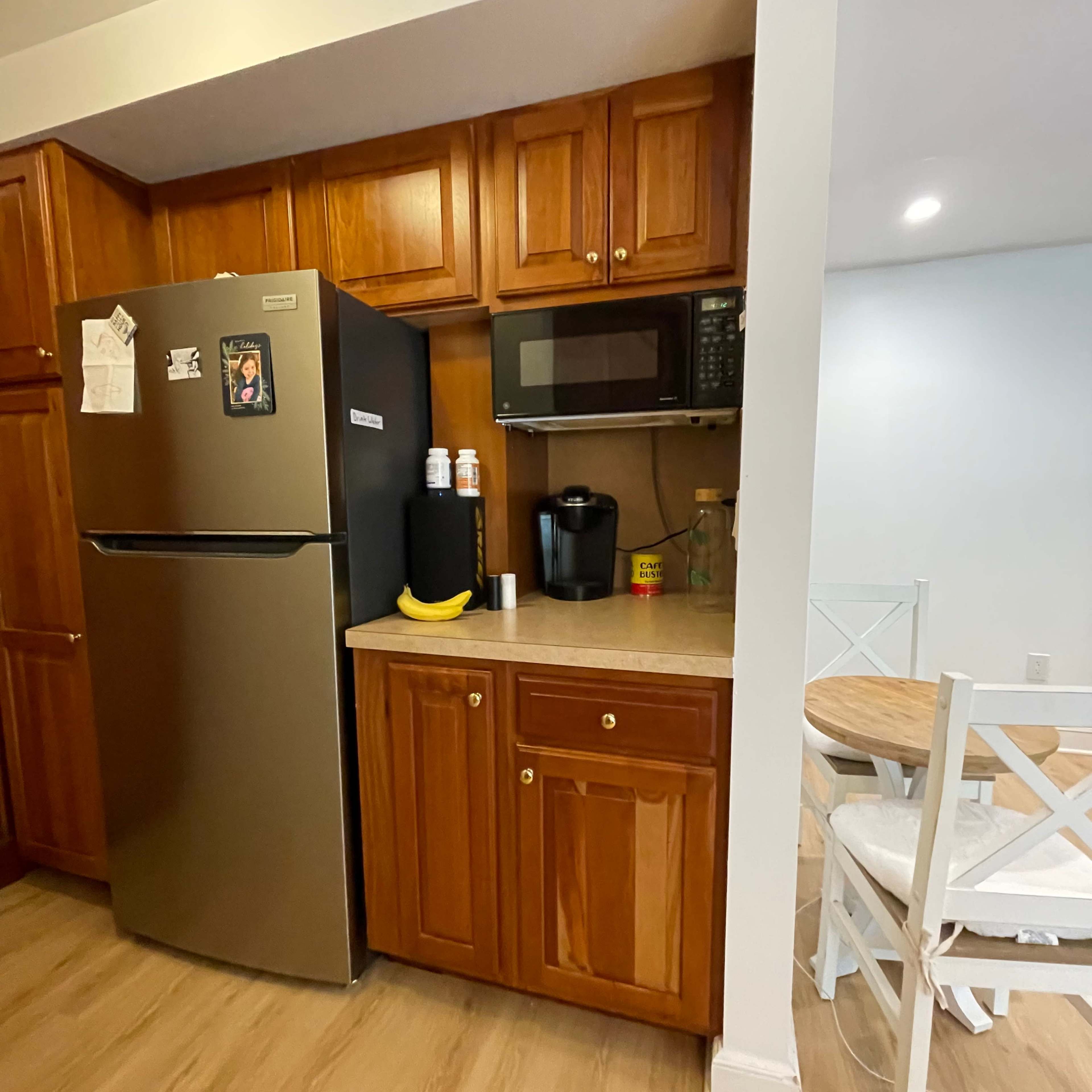 A kitchen area featuring a stainless steel refrigerator, a microwave, and a coffee maker, with wooden cabinets and a small dining table nearby.