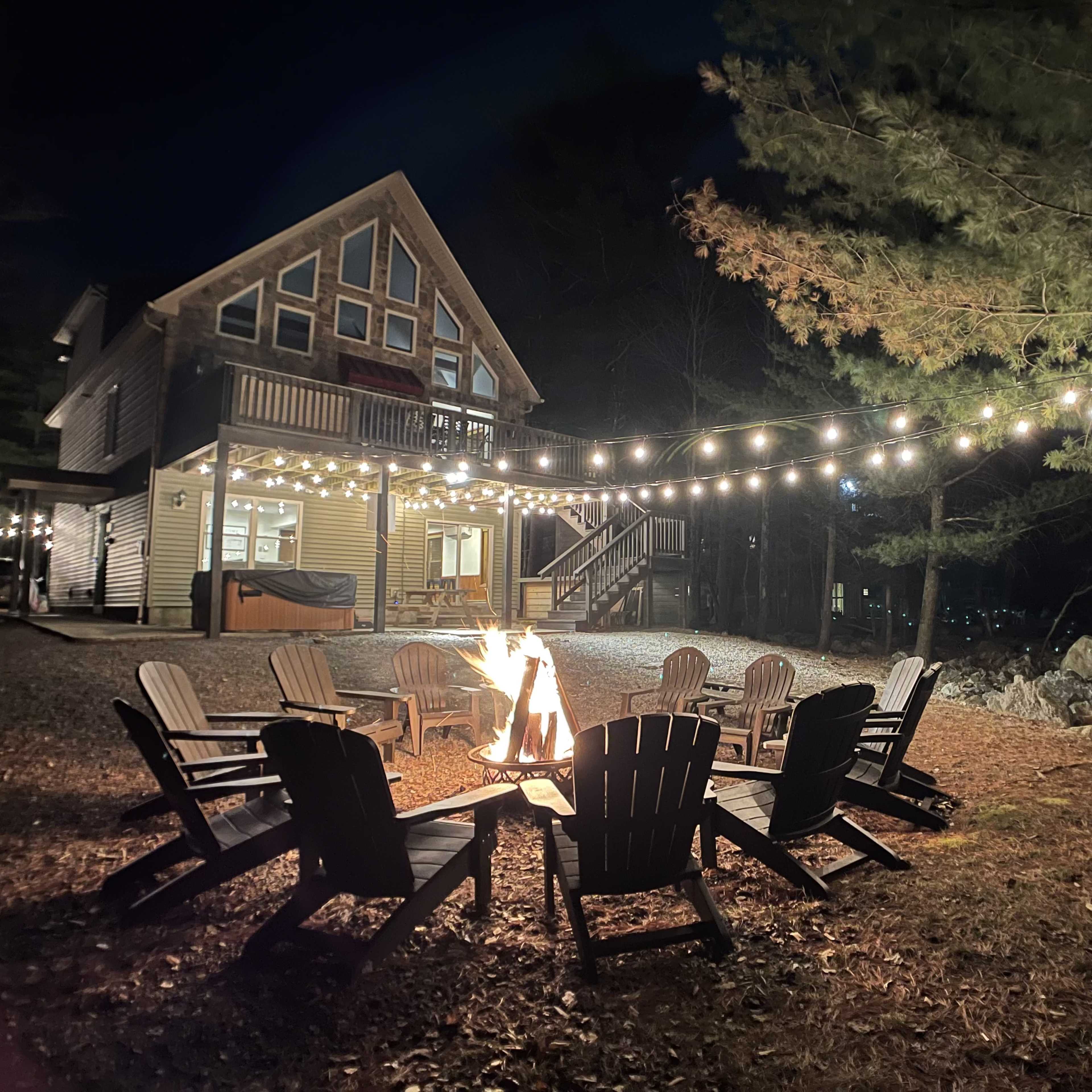 A fire pit surrounded by wooden chairs is set in front of a house illuminated by string lights at night, with trees in the background.