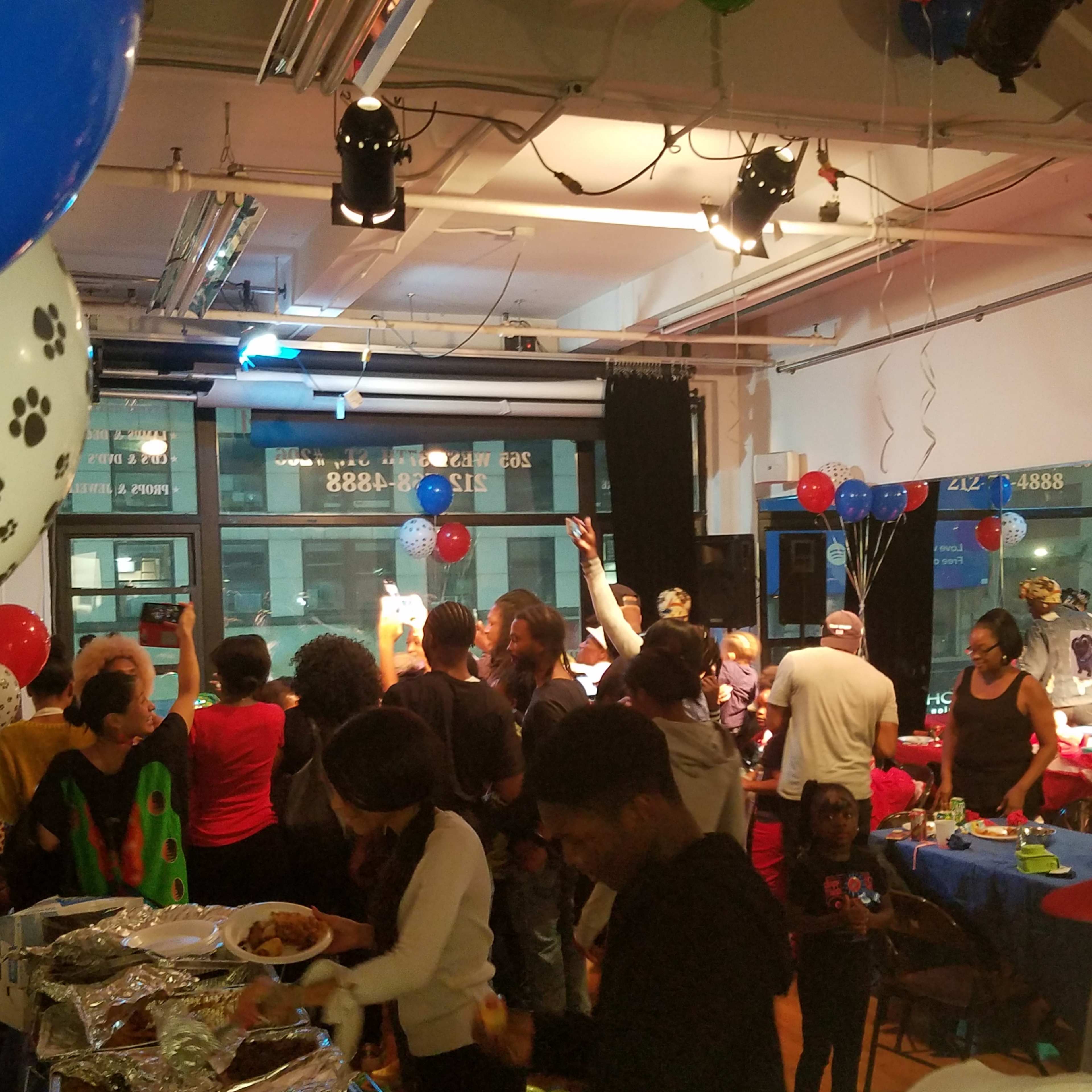 A large group of people gathers in a brightly lit event space decorated with balloons and tables of food for a celebration.