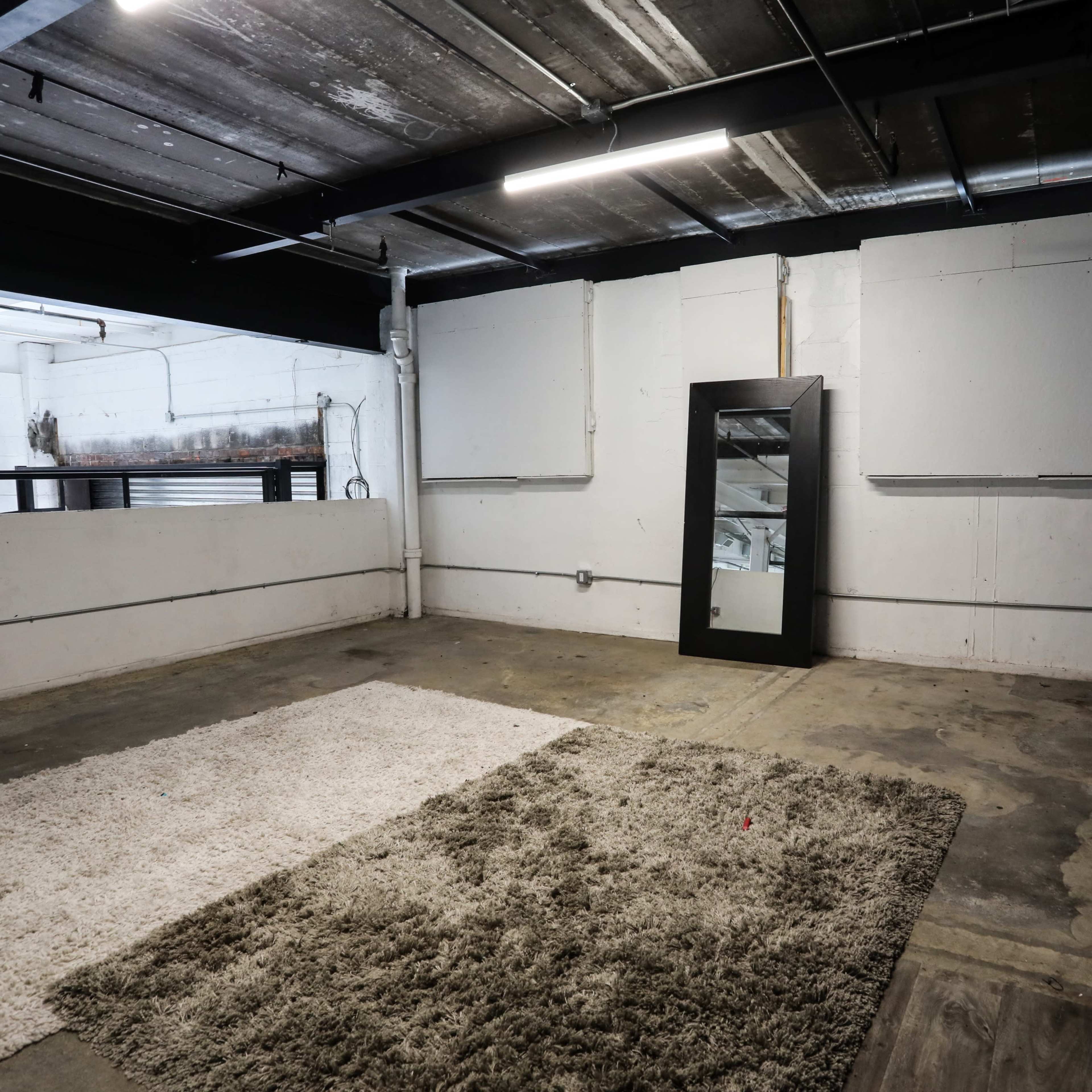 An empty industrial-style room with exposed concrete walls, a large mirror on one side, and a textured rug on the floor.