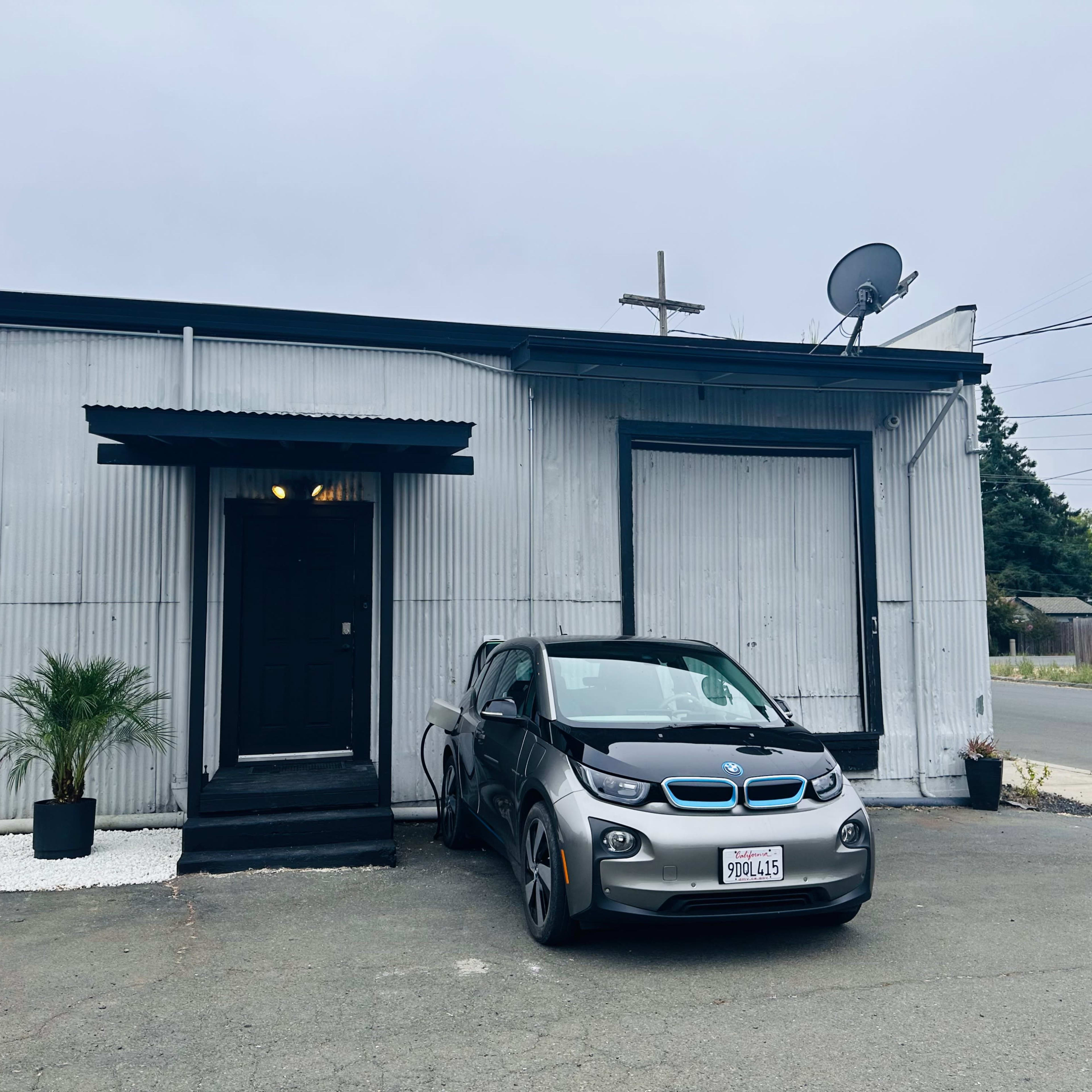 A grey BMW i3 is parked in front of a corrugated metal building with a black door and a satellite dish.
