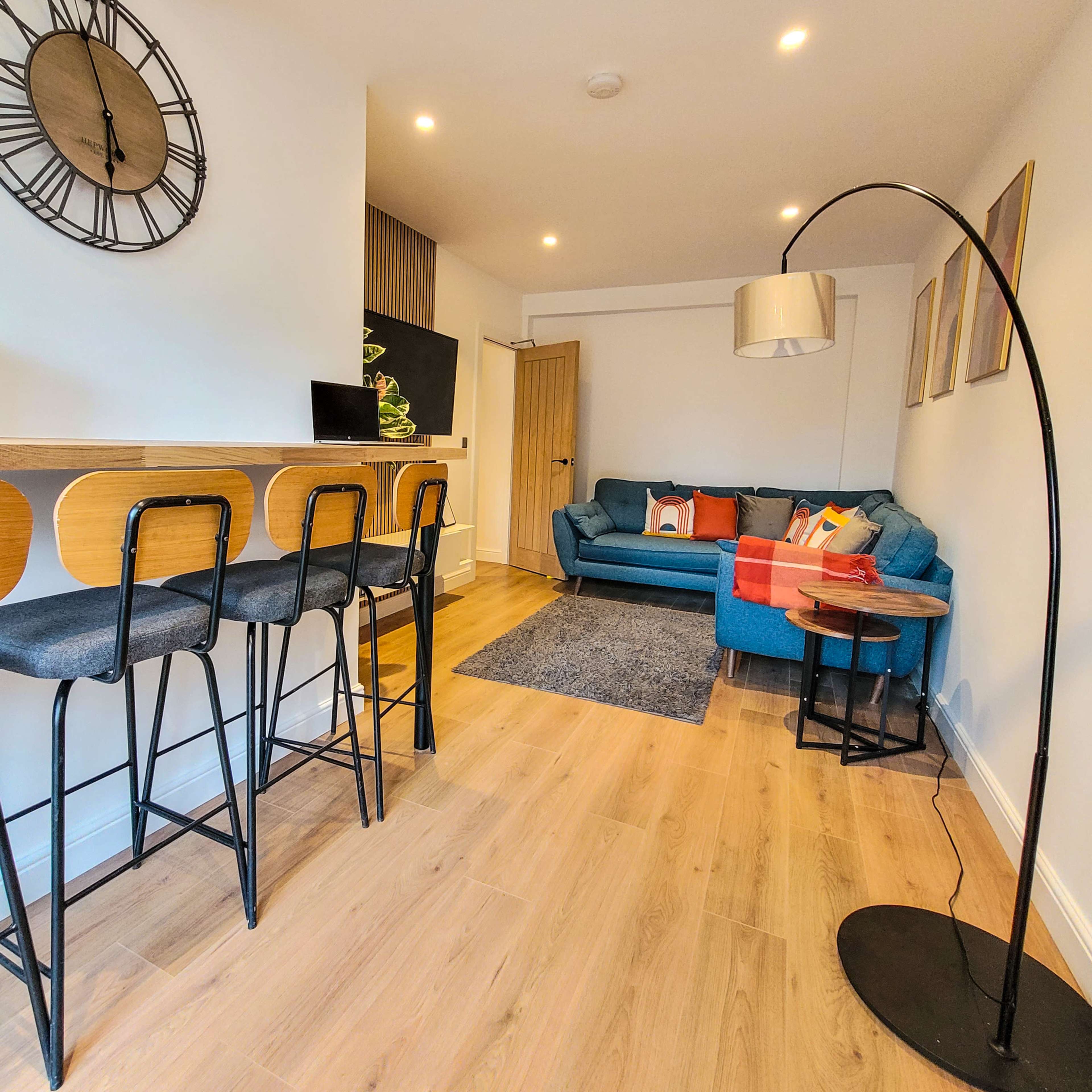 A modern open-plan living area featuring a seating arrangement with a blue couch, a small dining bar with stools, and a floor lamp.
