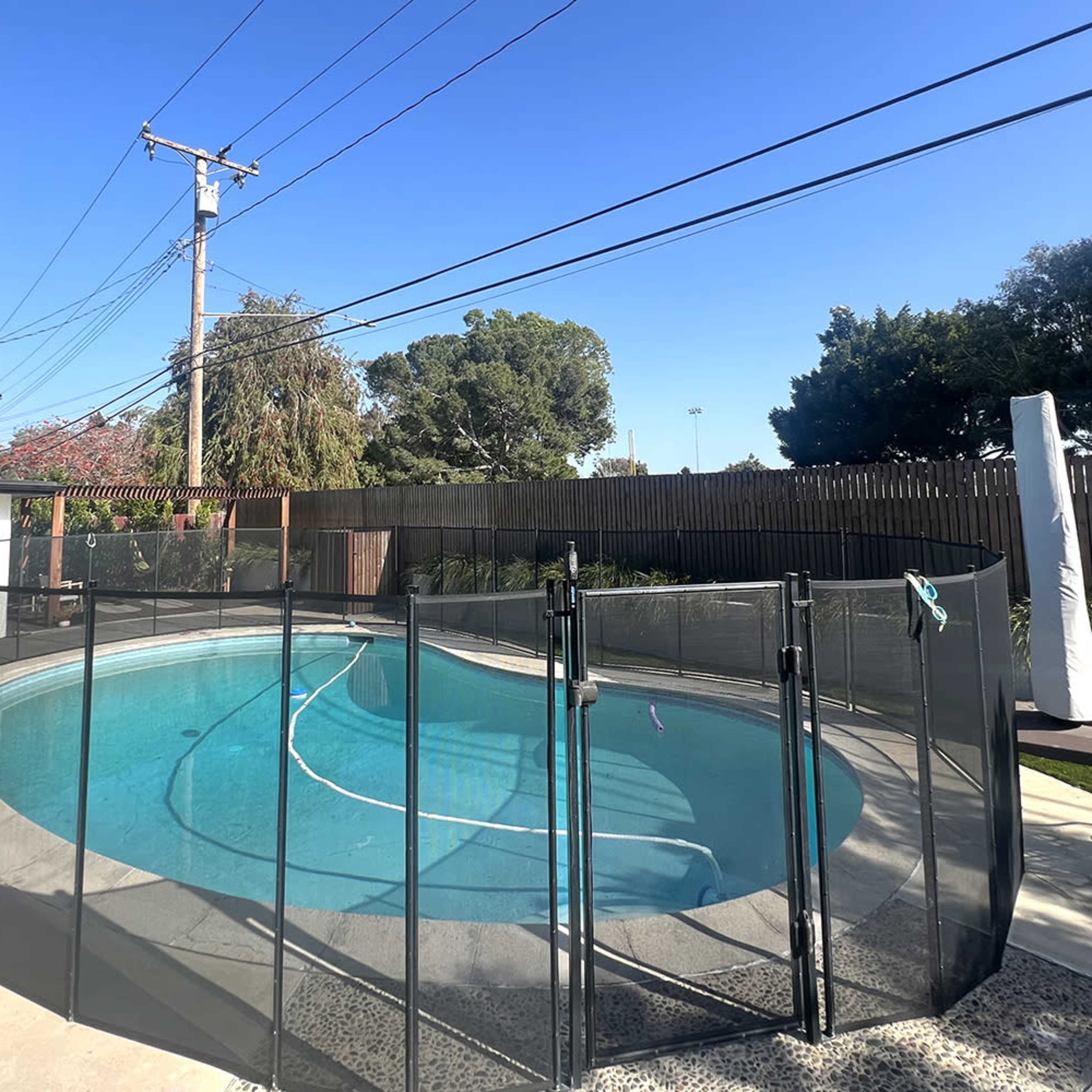 A swimming pool surrounded by a black safety fence, with a clear blue sky overhead and green foliage in the background.