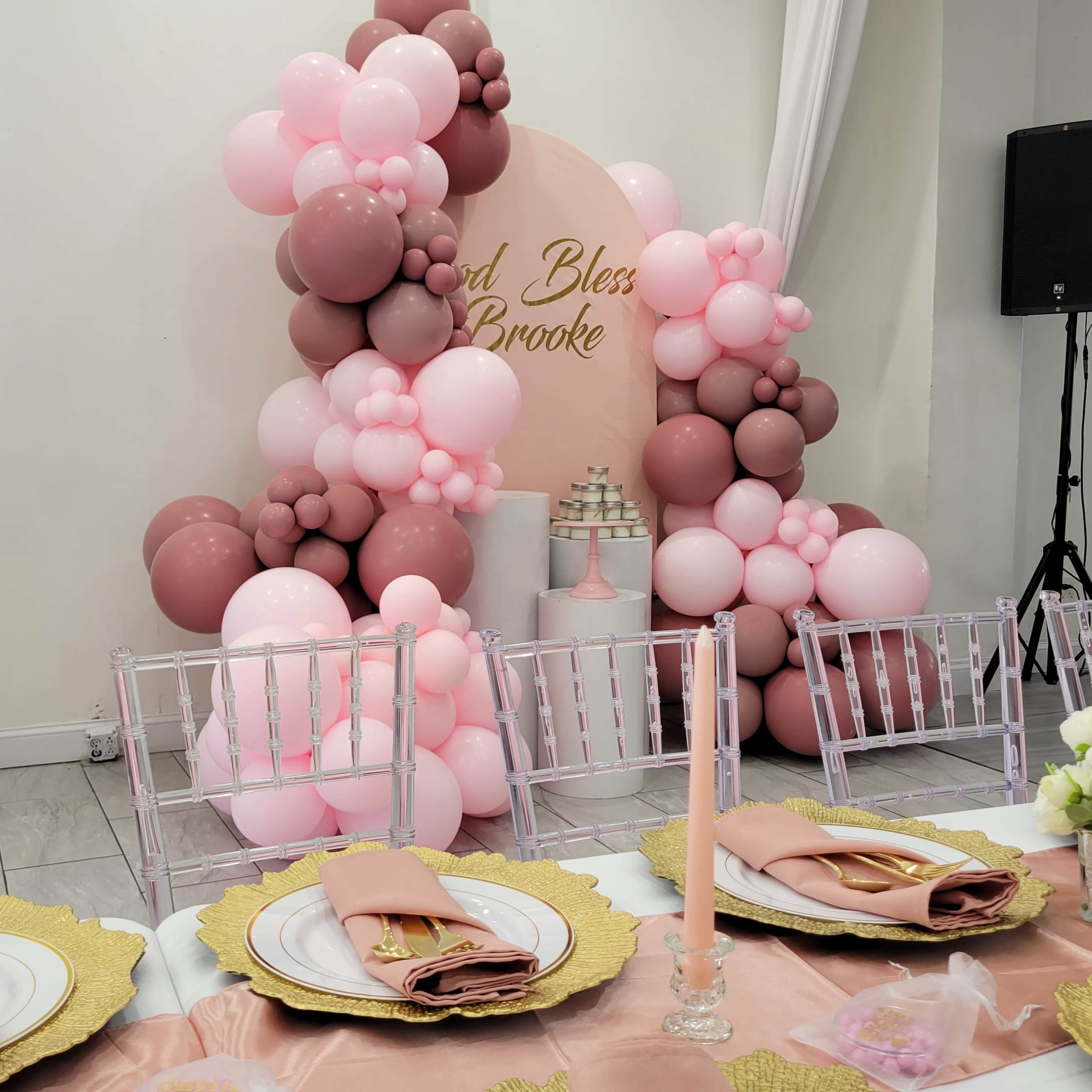 A table is set with elegant pink and gold decor in front of a backdrop of pink and brown balloons that reads "God Bless Brooke."