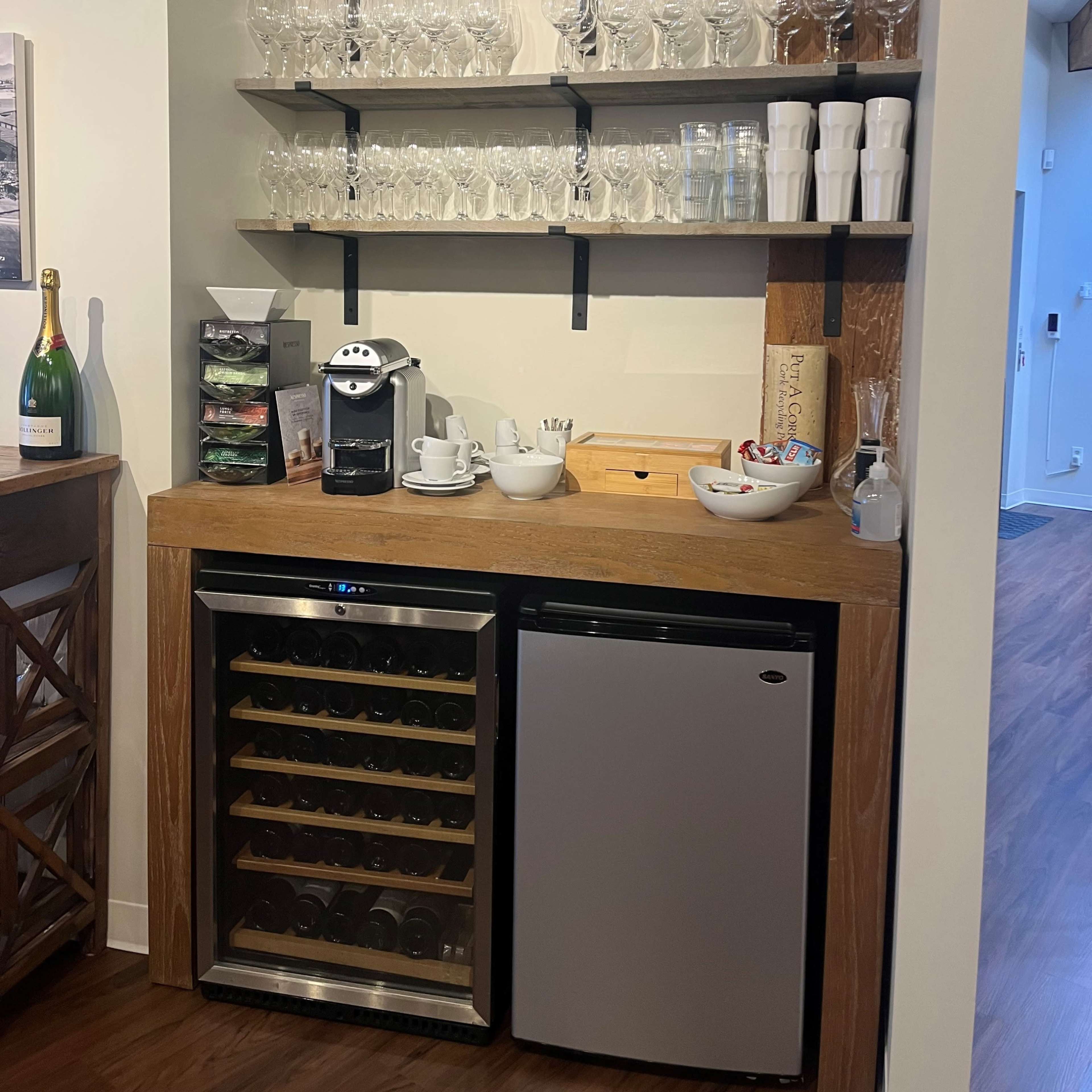 A wooden bar area with stacked glassware on shelves above a countertop featuring a coffee machine, dishware, and a small wine fridge.