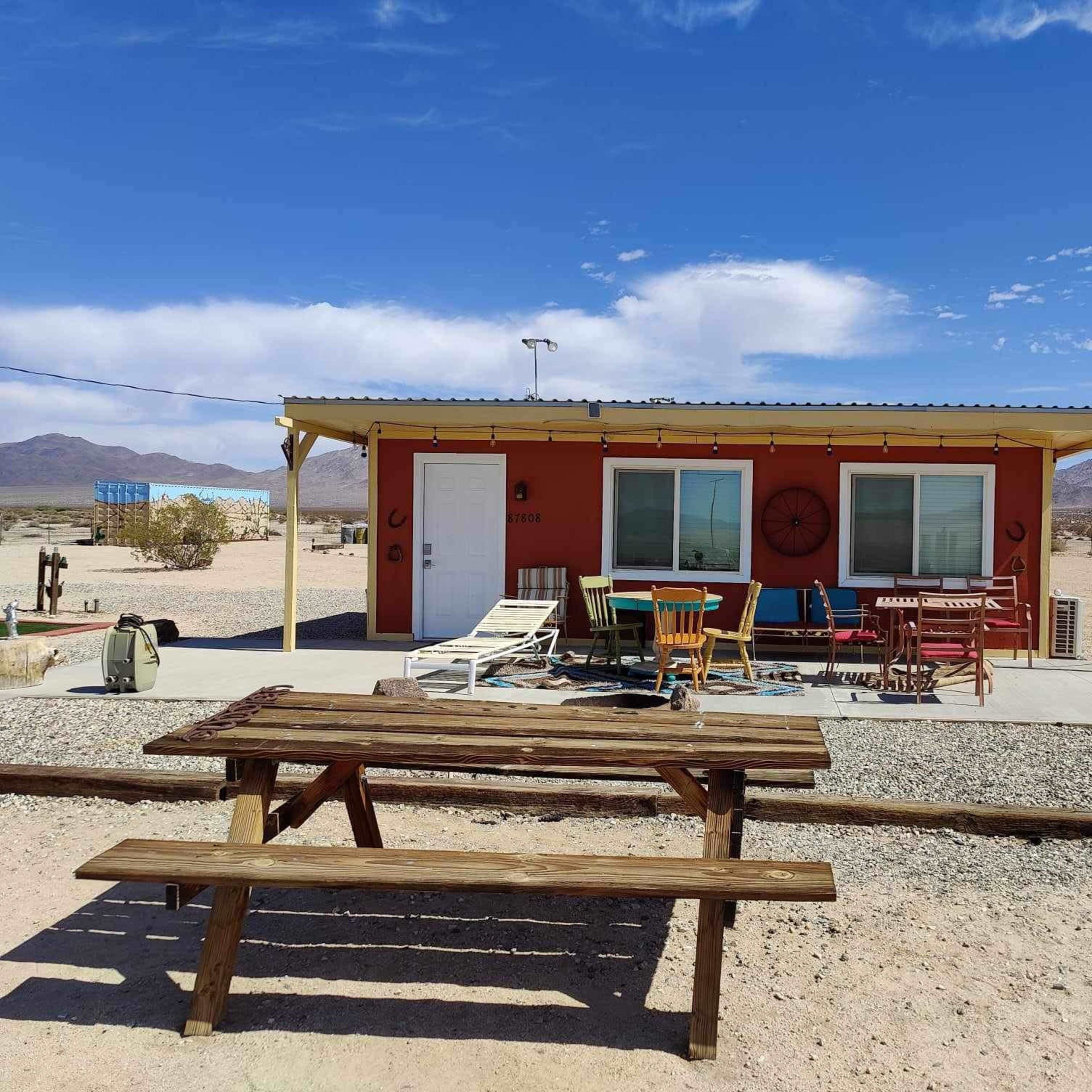 A colorful cabin with a front porch and outdoor seating is situated in a desert landscape with mountains in the background.