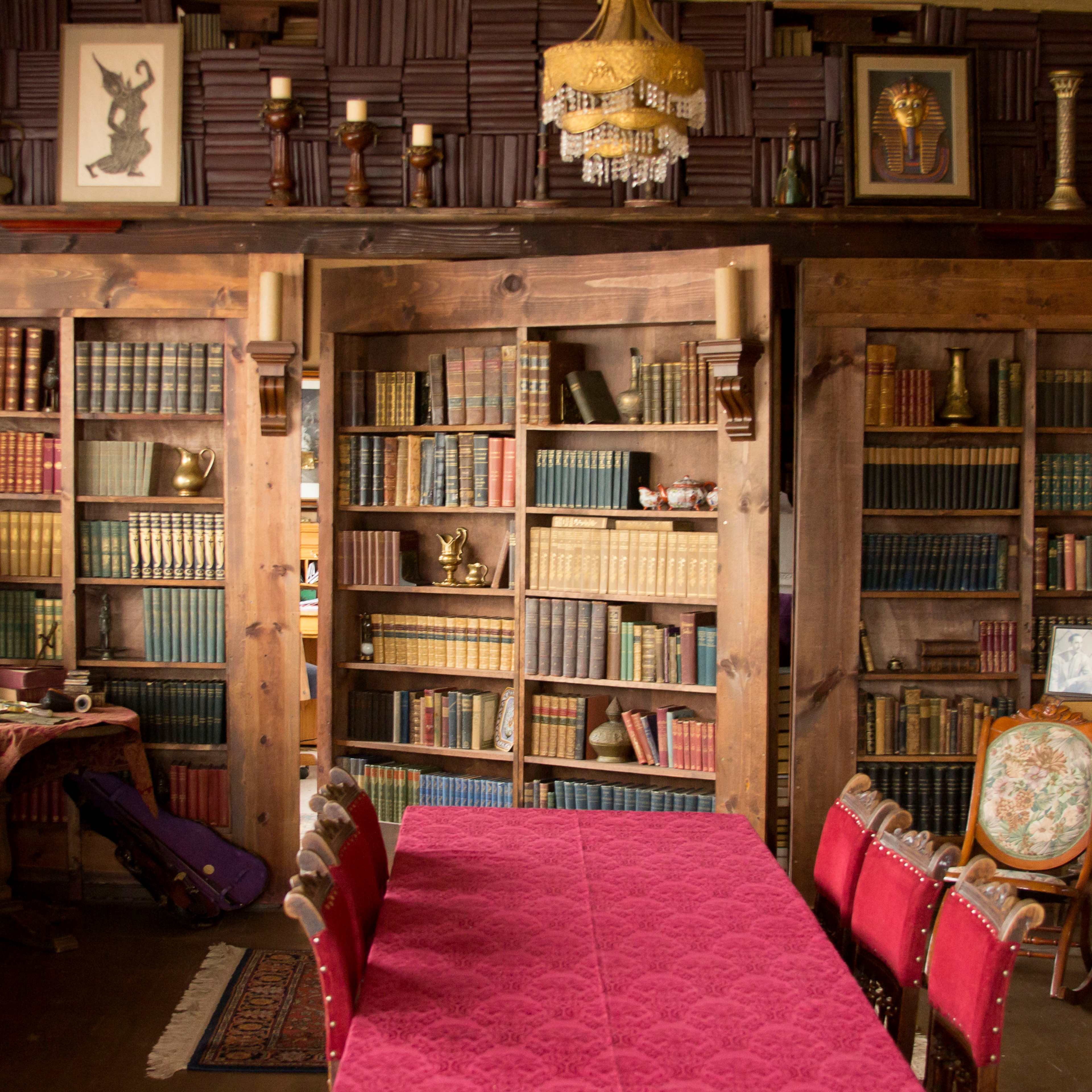 A room featuring a wooden bookshelf filled with books, framed art on the walls, and a large table with red chairs in front of it.