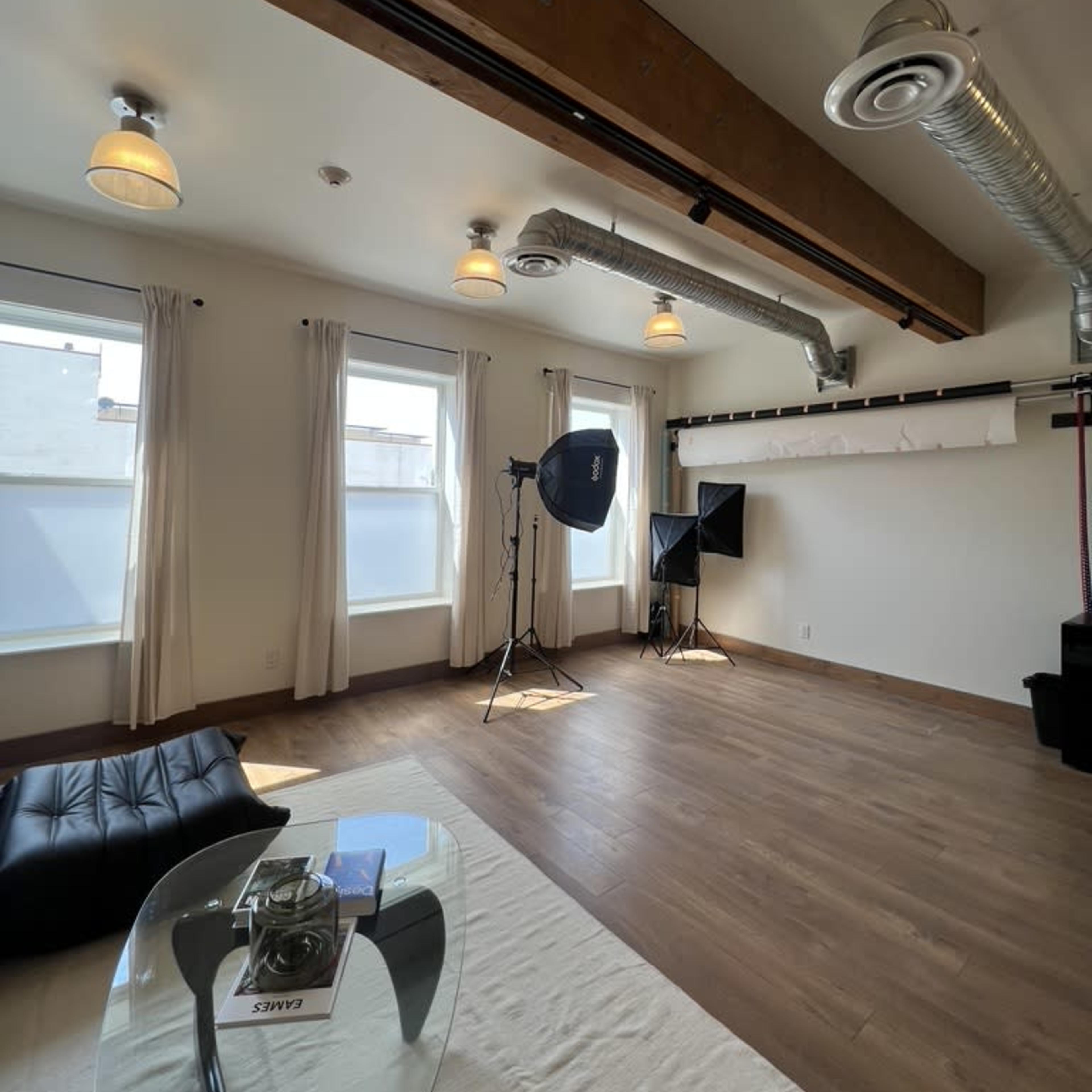 A spacious room with wooden floors, large windows, two light stands with softboxes, and minimal furnishings including a black couch and a glass table.