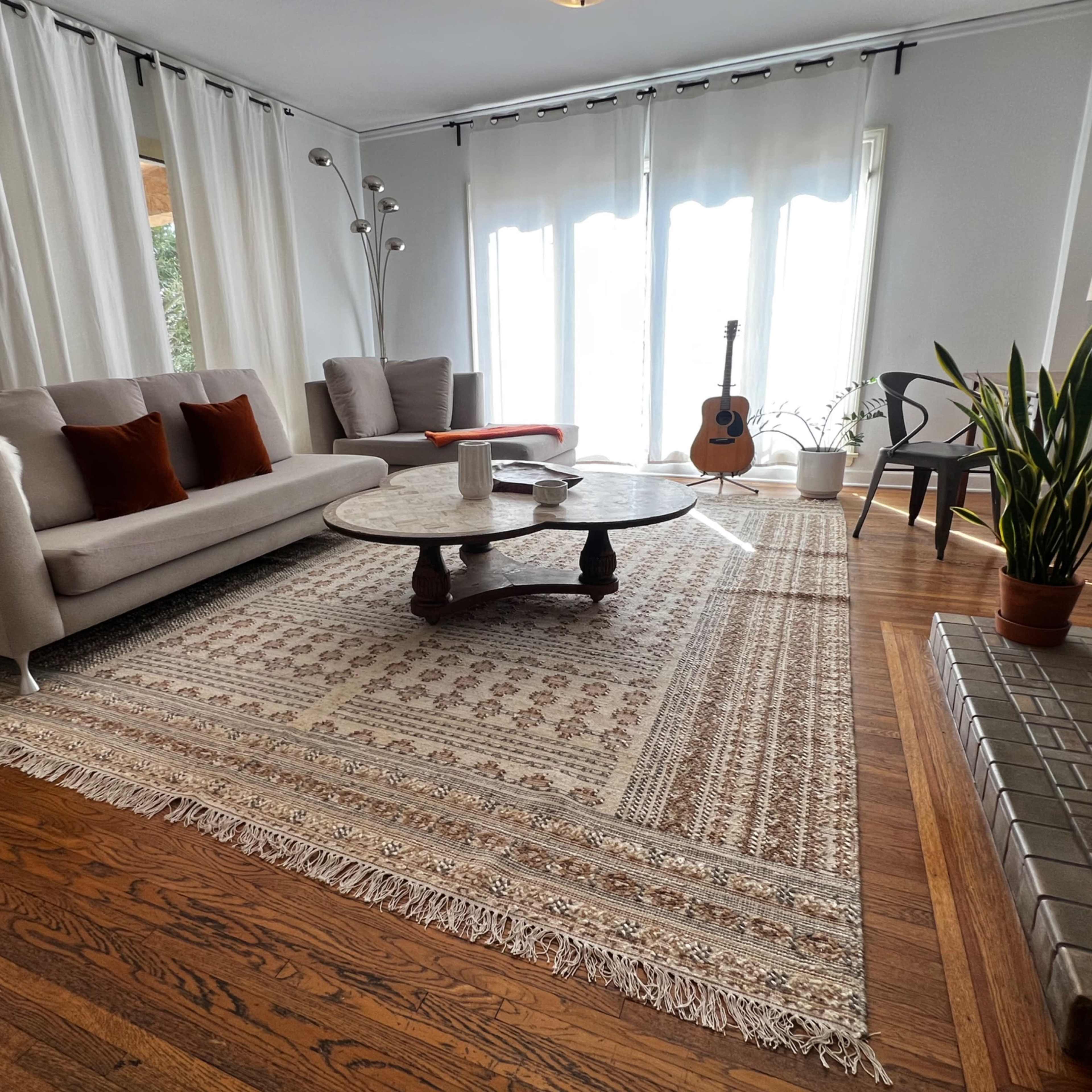 A light-filled living room with a sofa, a round coffee table, a guitar, and plants, all situated on a patterned area rug.