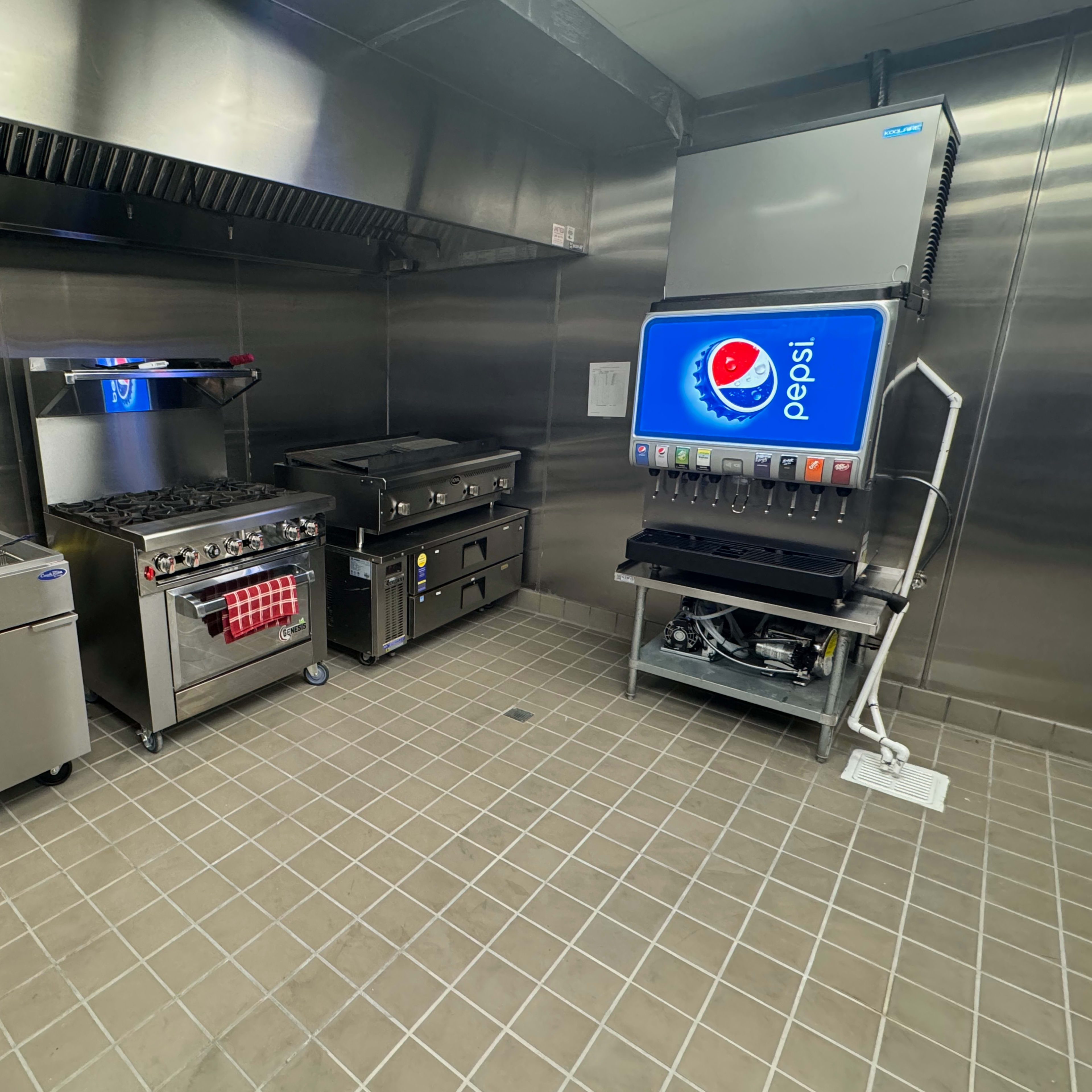 A commercial kitchen with stainless steel equipment, including a gas range, a flat top grill, a beverage dispenser, and a storage unit along tiled flooring.