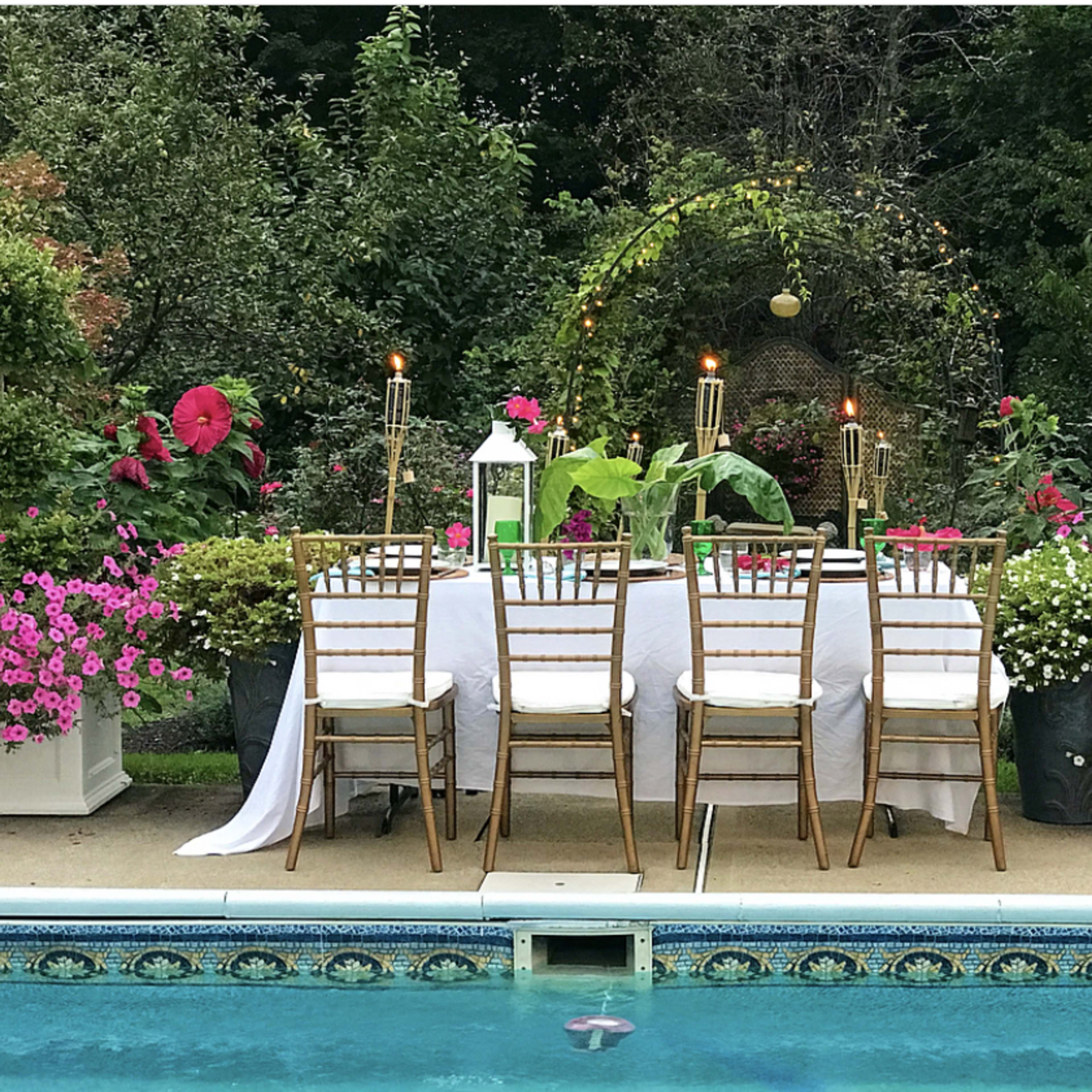 A beautifully set dining table with gold chairs is arranged by a swimming pool surrounded by vibrant flowers.