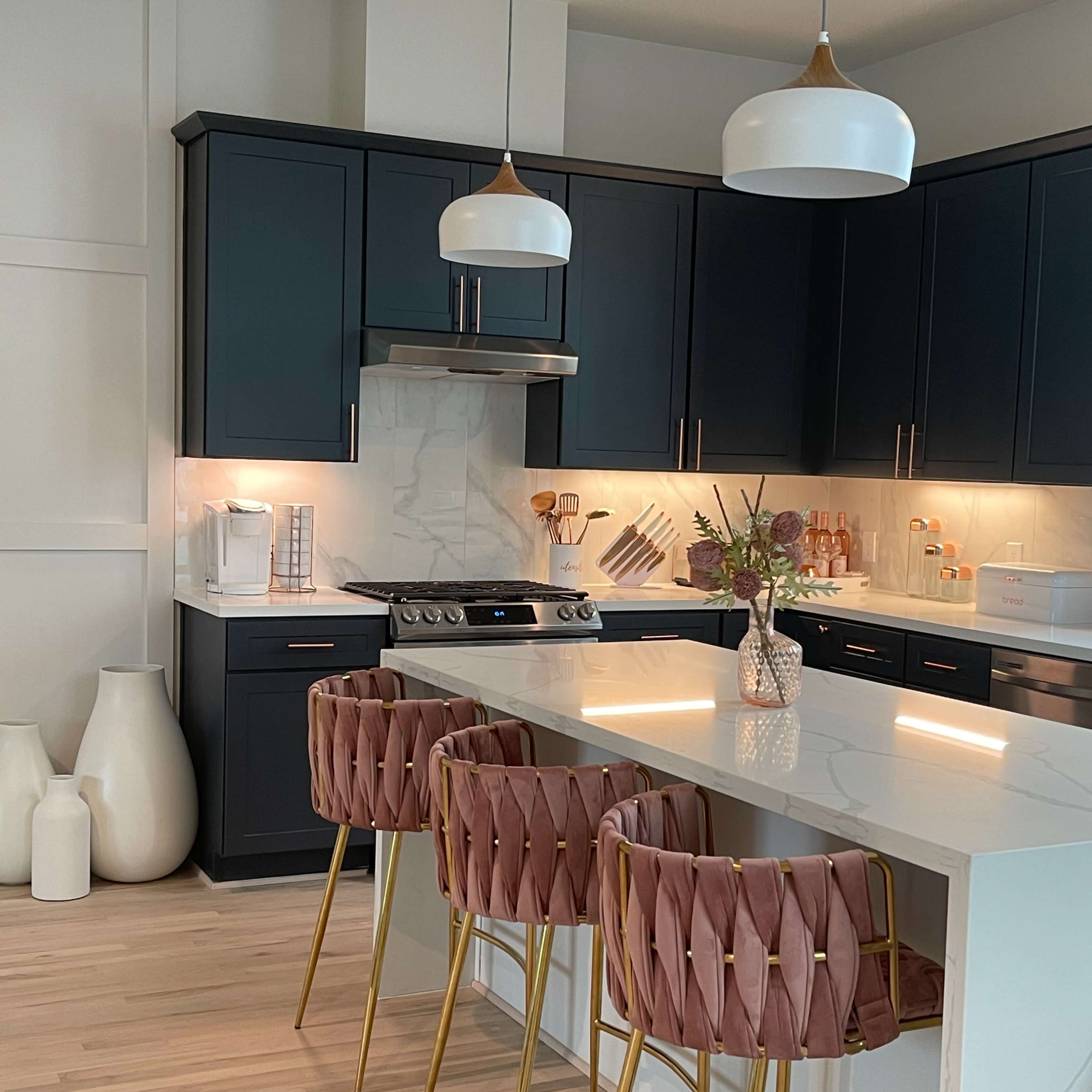 A modern kitchen with dark cabinetry, white countertops, and three pink and gold barstools at a central island.