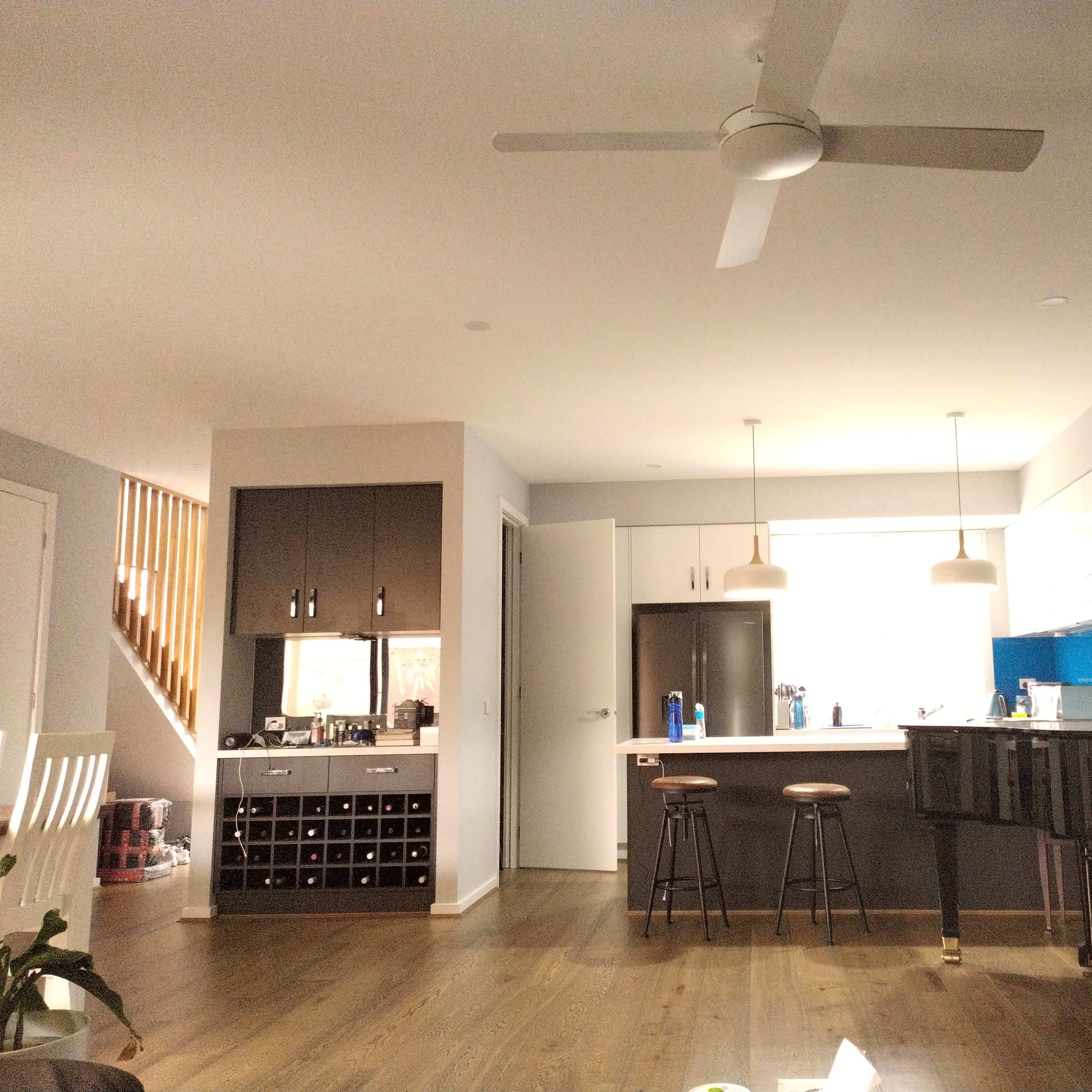 A modern kitchen and living area featuring a piano, bar stools, and a wine rack, with a ceiling fan above.