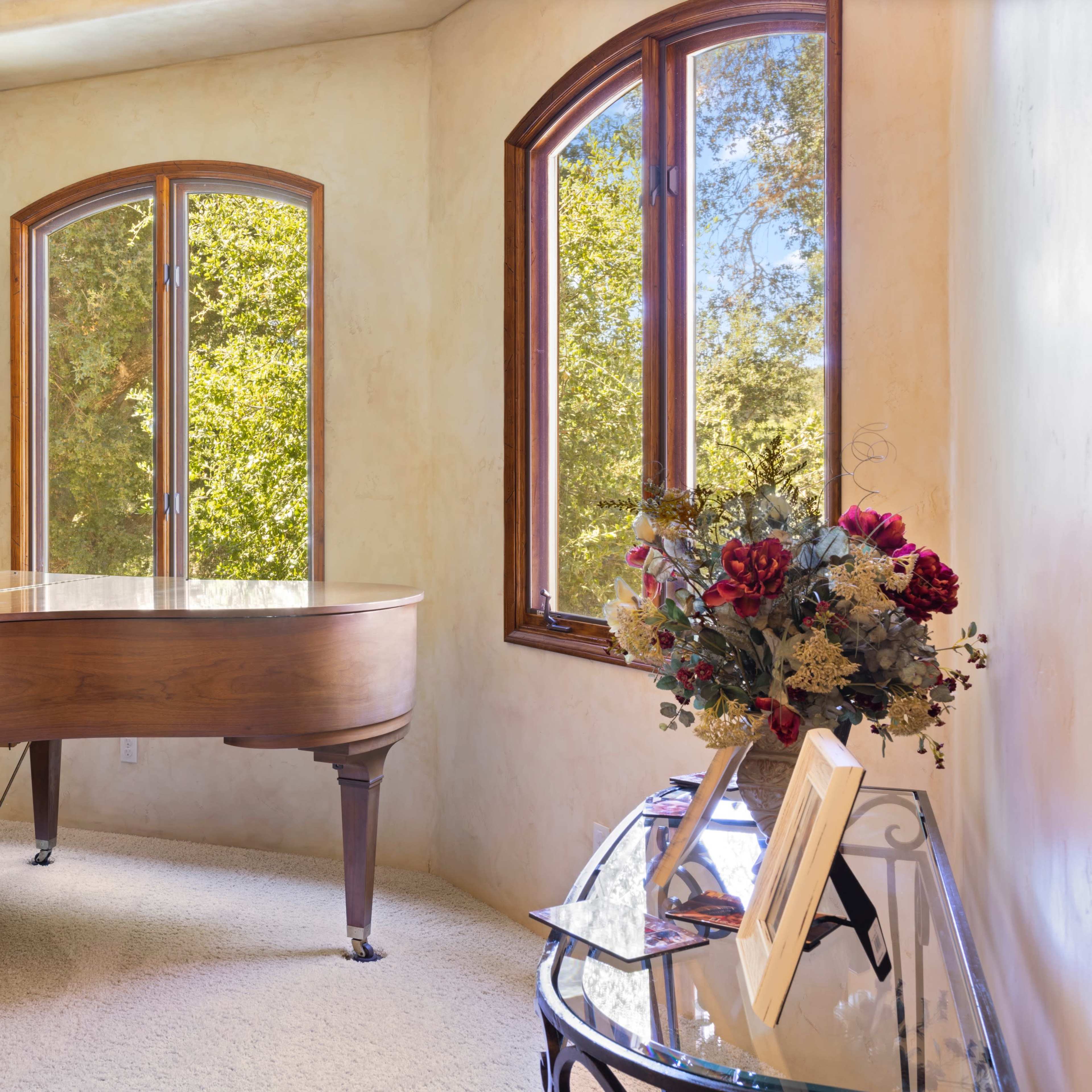 A grand piano is positioned in a sunlit corner with large windows overlooking greenery, accompanied by a decorative table adorned with flowers and books.