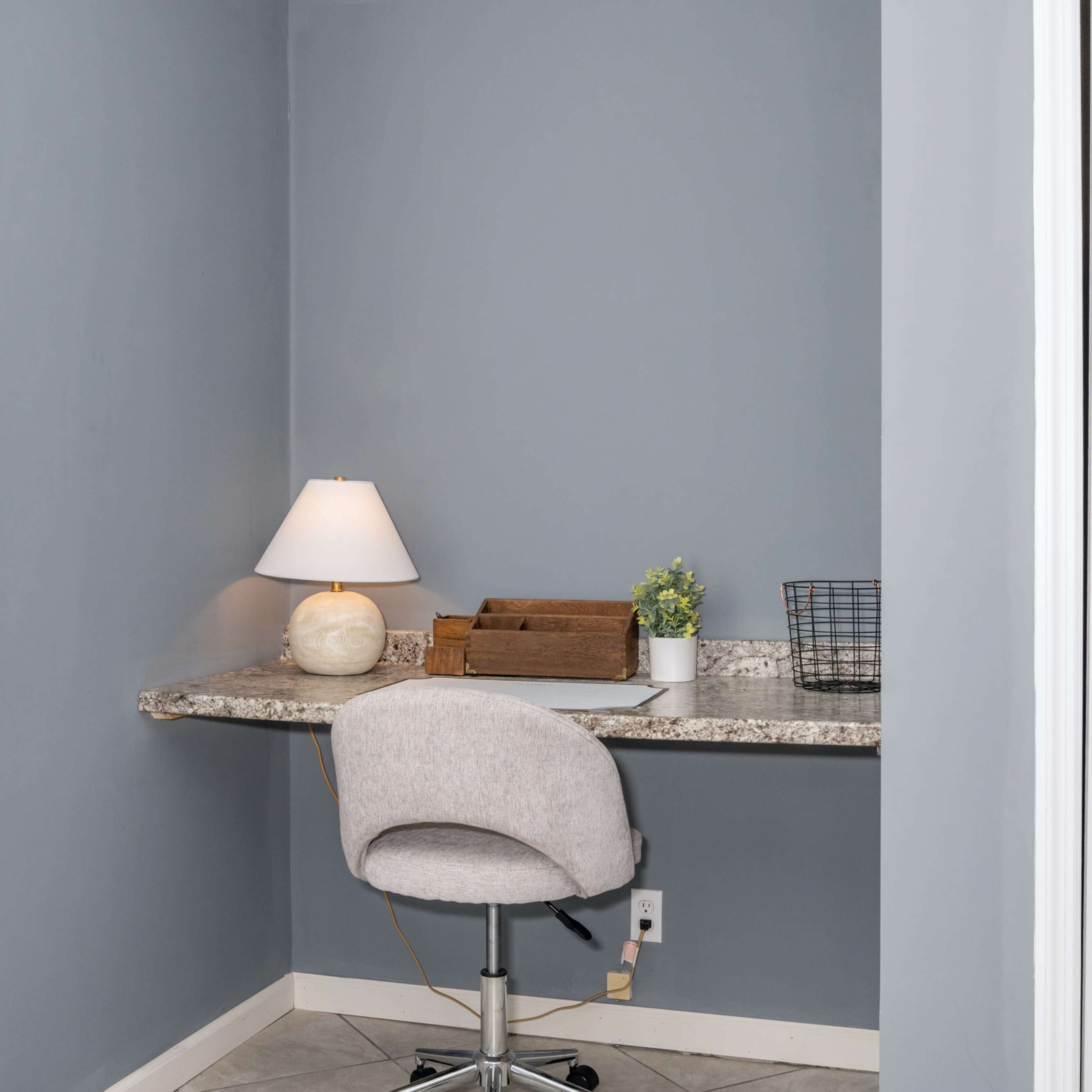 A small home workspace with a gray wall, a desk with a lamp, a wooden organizer, a potted plant, and a mesh basket.