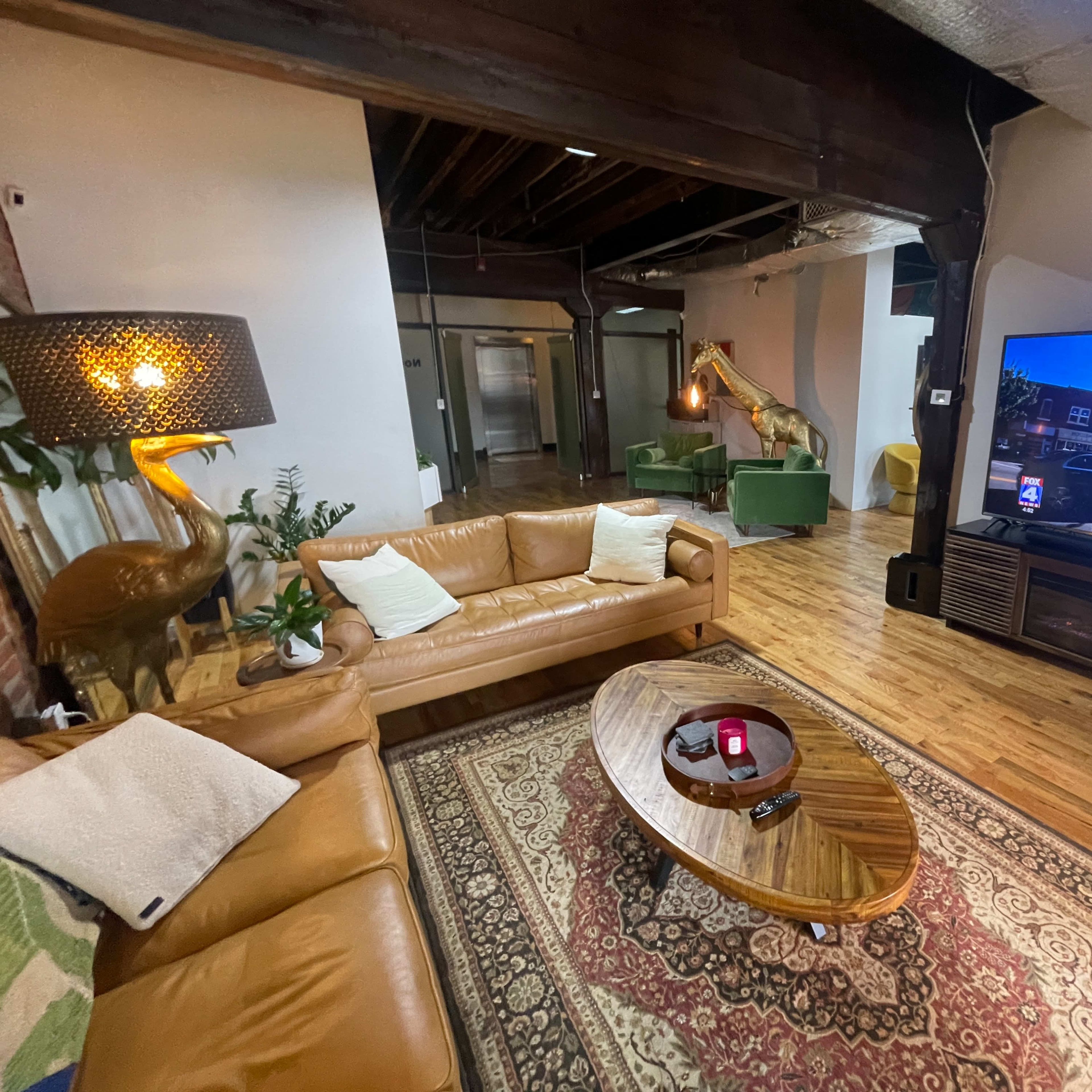 A cozy living room with a brown leather sofa, a round wooden coffee table, a large TV mounted on the wall, and various indoor plants.