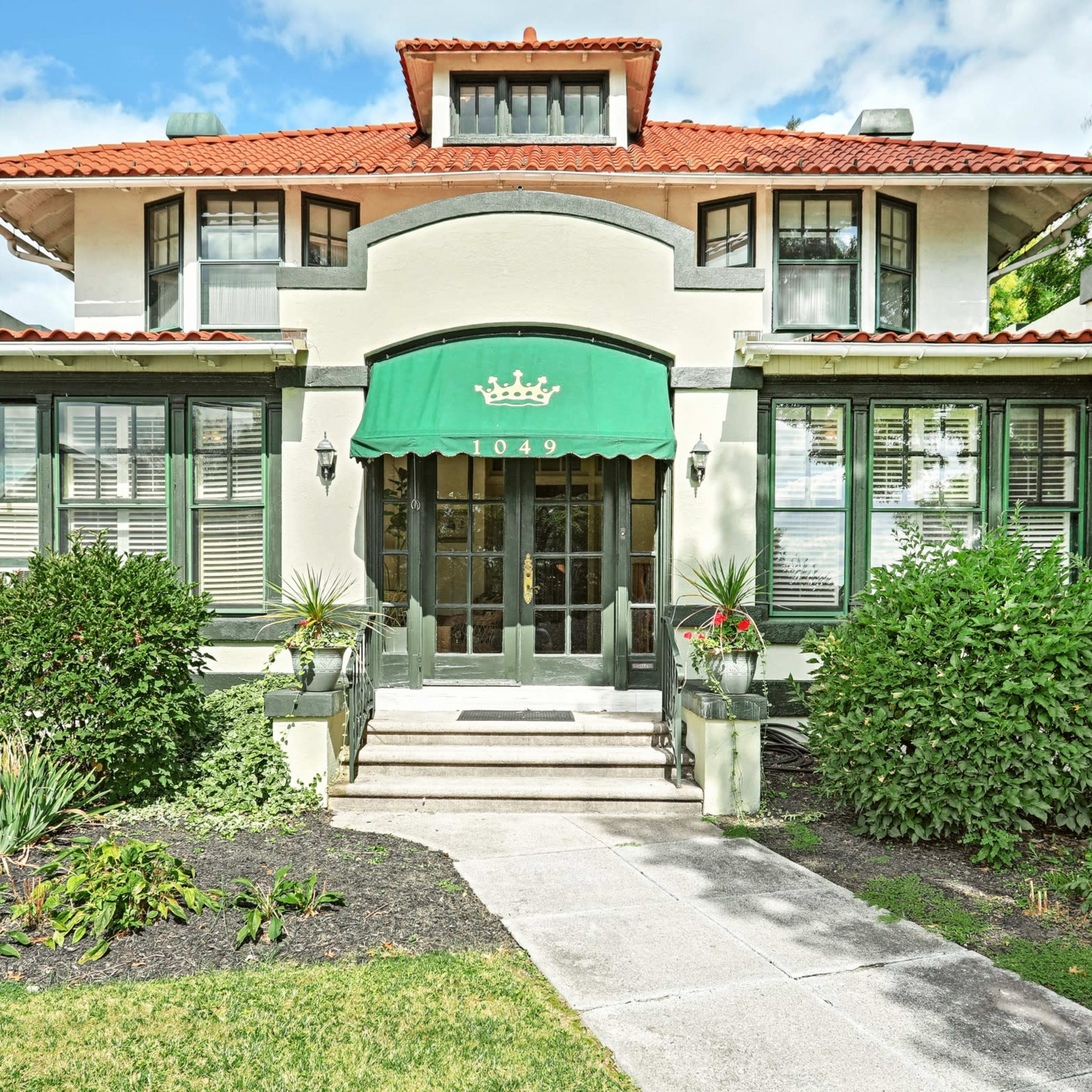 A two-story house with a red tile roof and a green awning, featuring symmetrical windows and well-maintained landscaping, stands prominently at the address 1049.