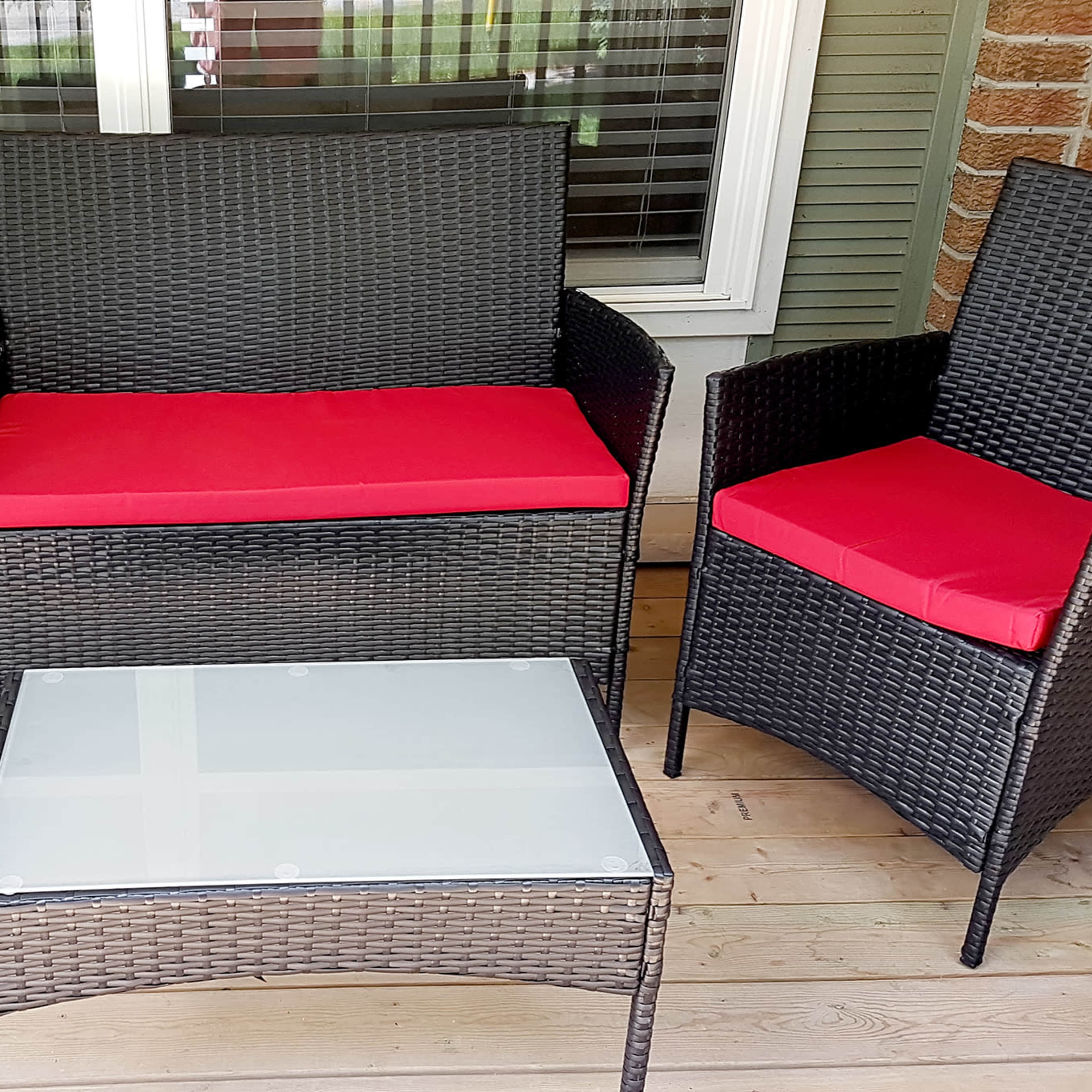 A seating area with two black wicker armchairs and a matching loveseat, all featuring red cushions, arranged around a glass-top coffee table on a wooden porch.