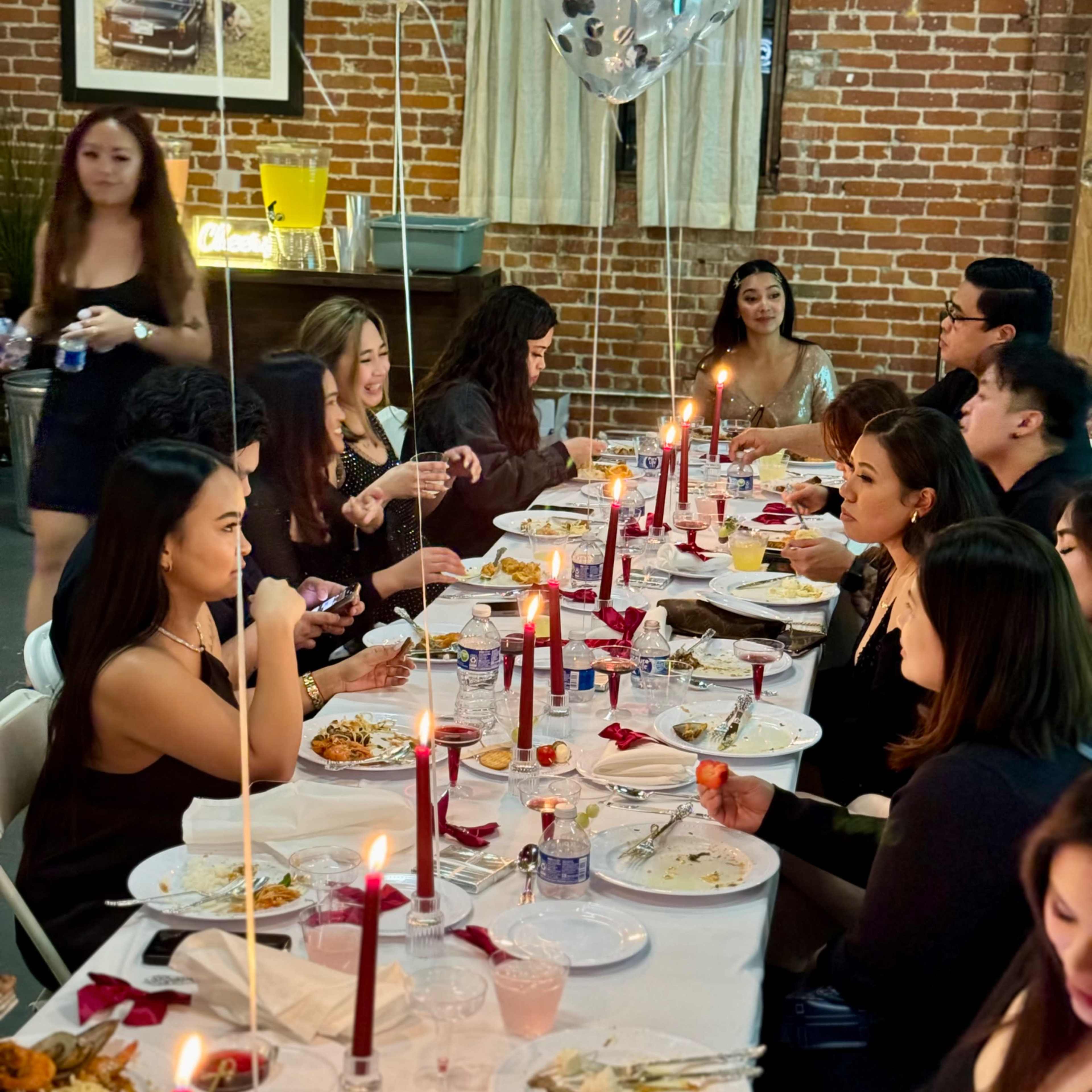 A long table is set for a dinner party, lined with candles and red balloons, while guests engage in conversation, enjoying their meals.