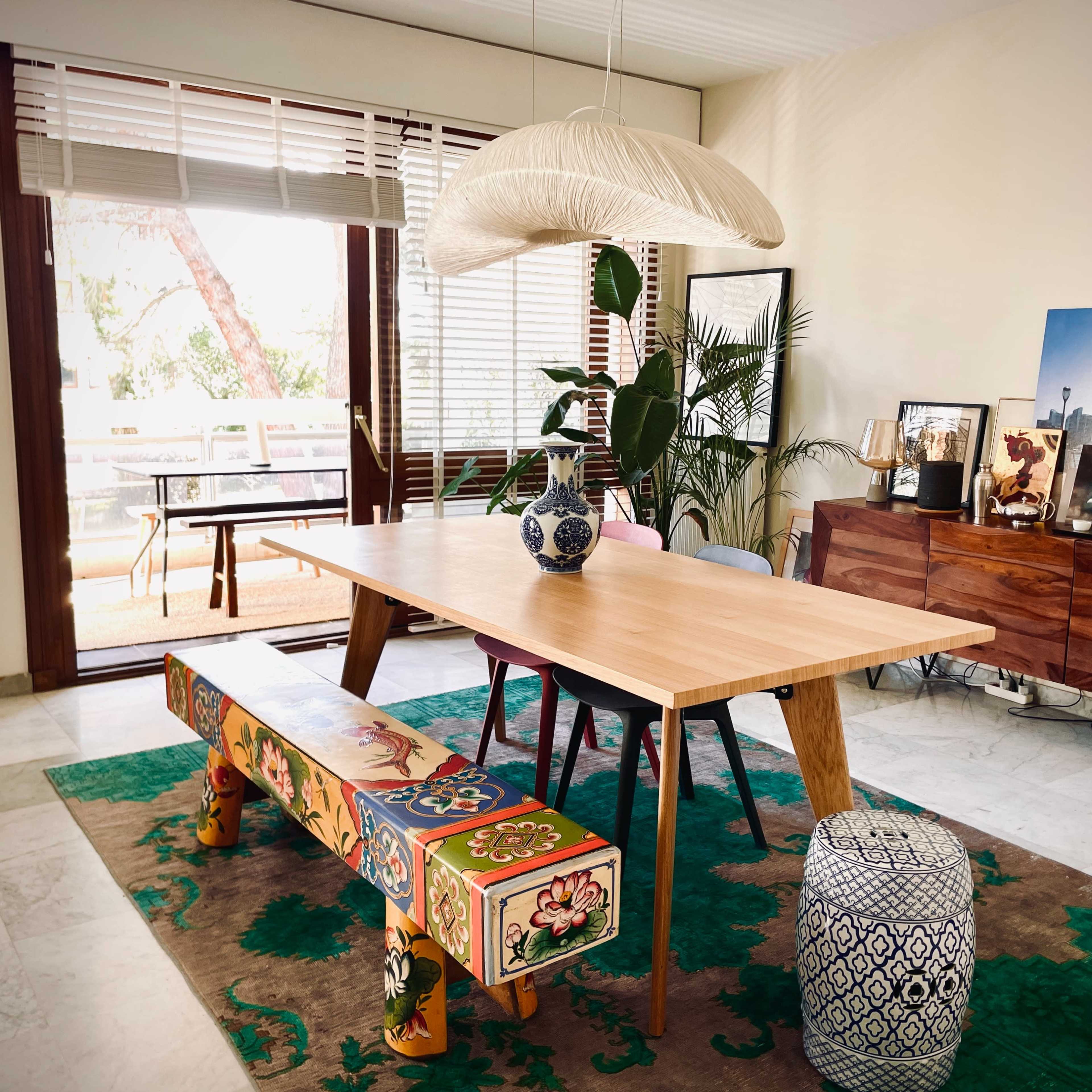 A dining room features a wooden table with a decorative vase, surrounded by colorful stools and a rug, against a backdrop of large windows and a plant.