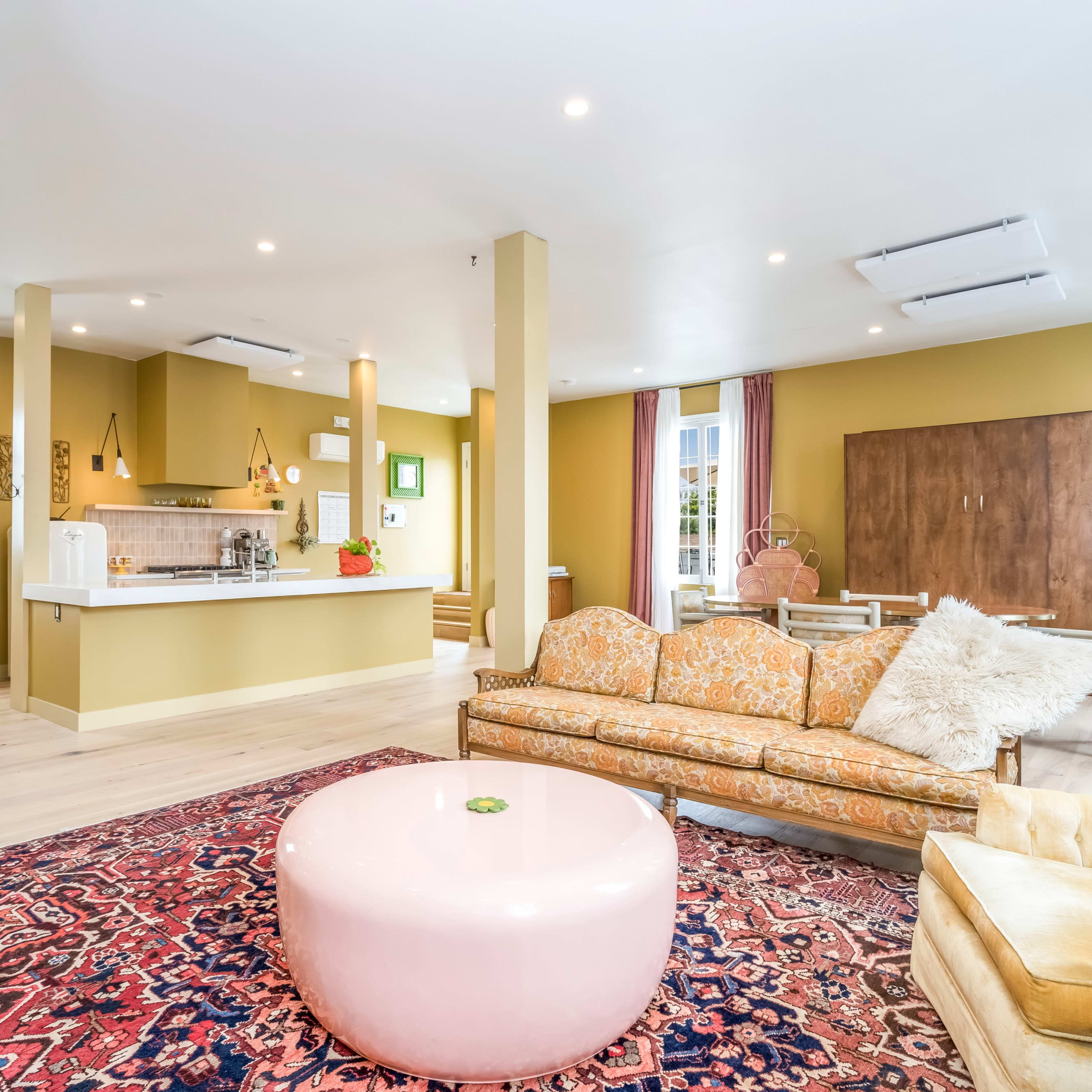 A spacious living area featuring a vintage-style sofa, a round pink coffee table, and a dining area with a large window, all set against a warm, olive-green wall.