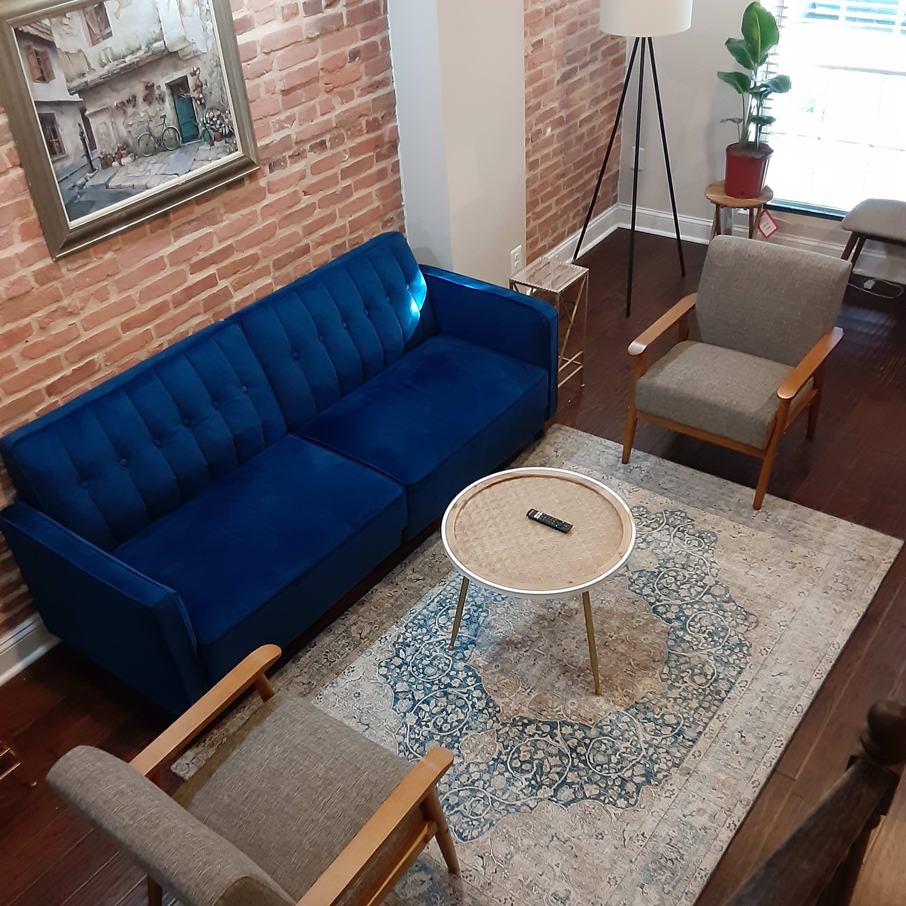 A living room with a blue sofa, two gray armchairs, a round coffee table, and a patterned rug, all set against a backdrop of exposed brick walls.