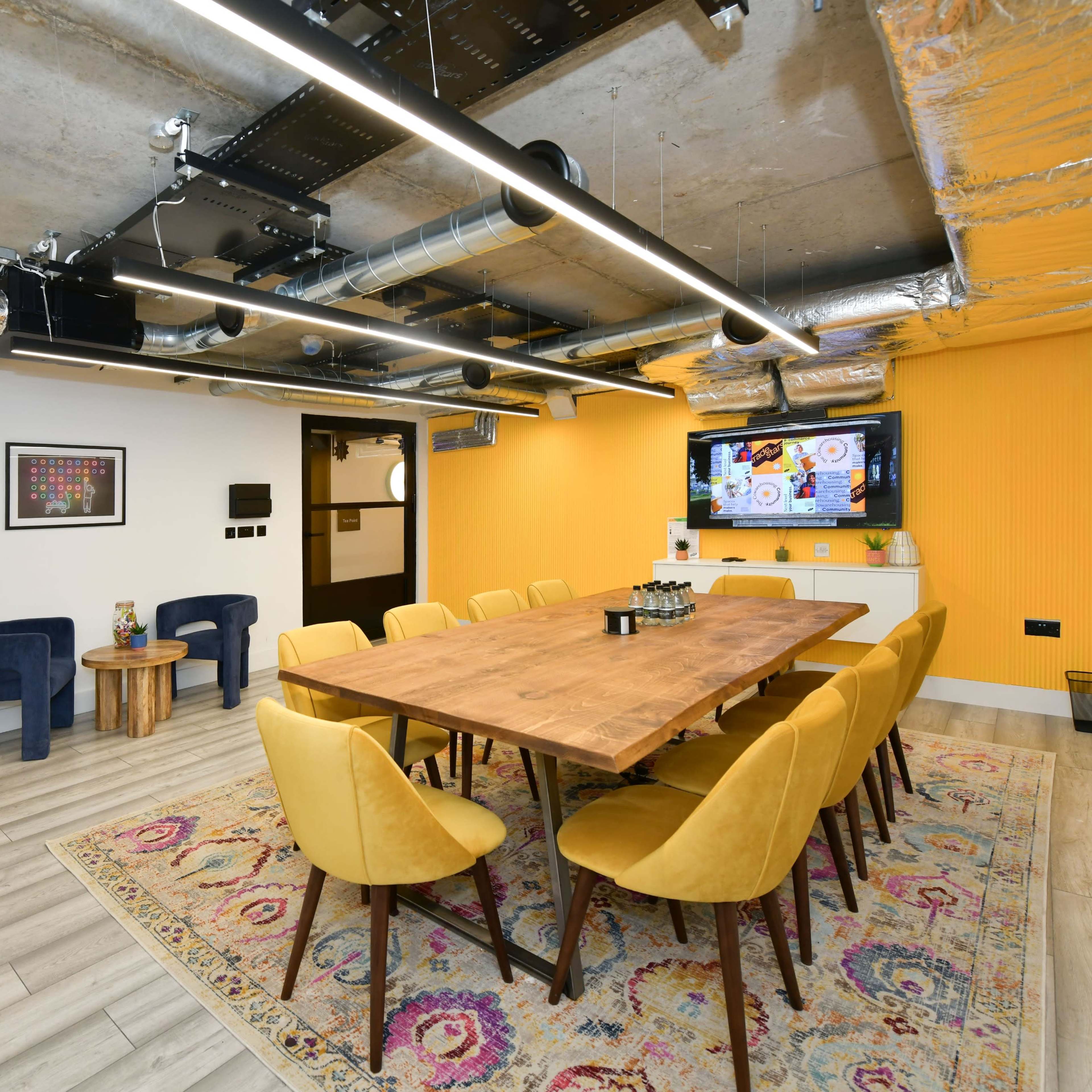 A modern conference room features a long wooden table surrounded by yellow chairs, with an orange accent wall and large screen display.