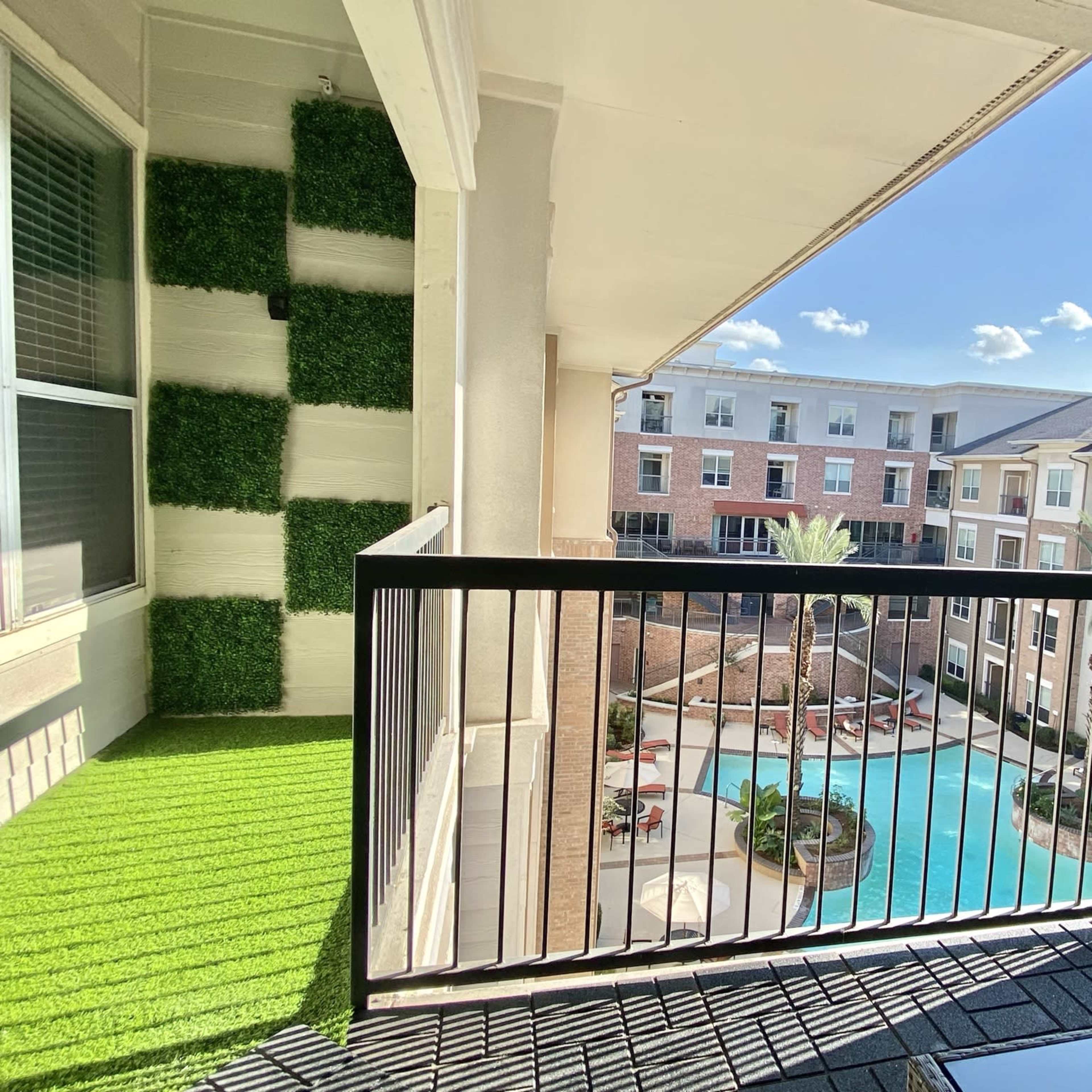 A balcony overlooking a swimming pool surrounded by apartment buildings, with artificial grass and greenery decor on the sides.