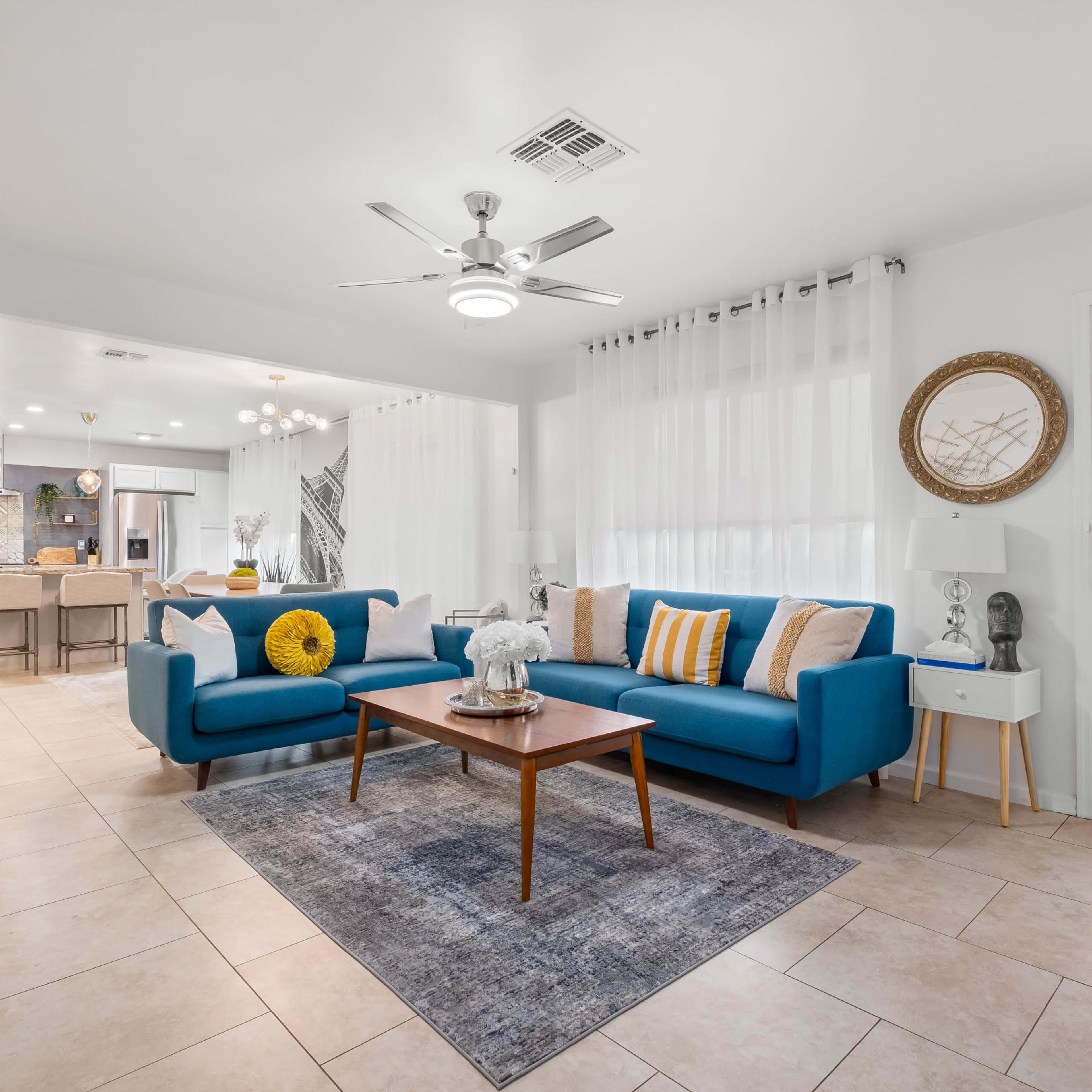 A modern living room features a blue sofa and armchair arranged around a wooden coffee table on a tiled floor, with an open kitchen visible in the background.