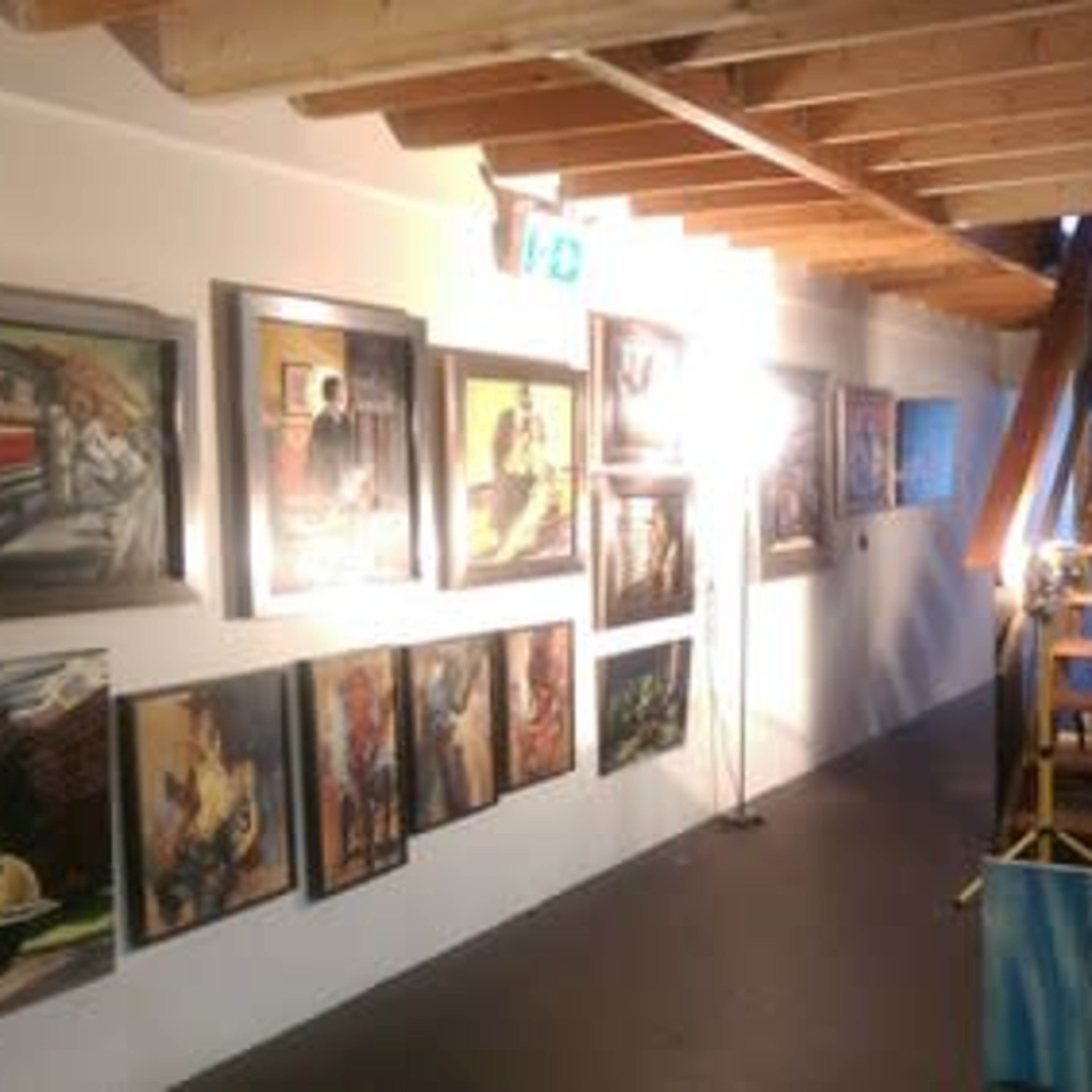 An art gallery hallway with framed paintings on the walls and a wooden staircase leading to a higher level.