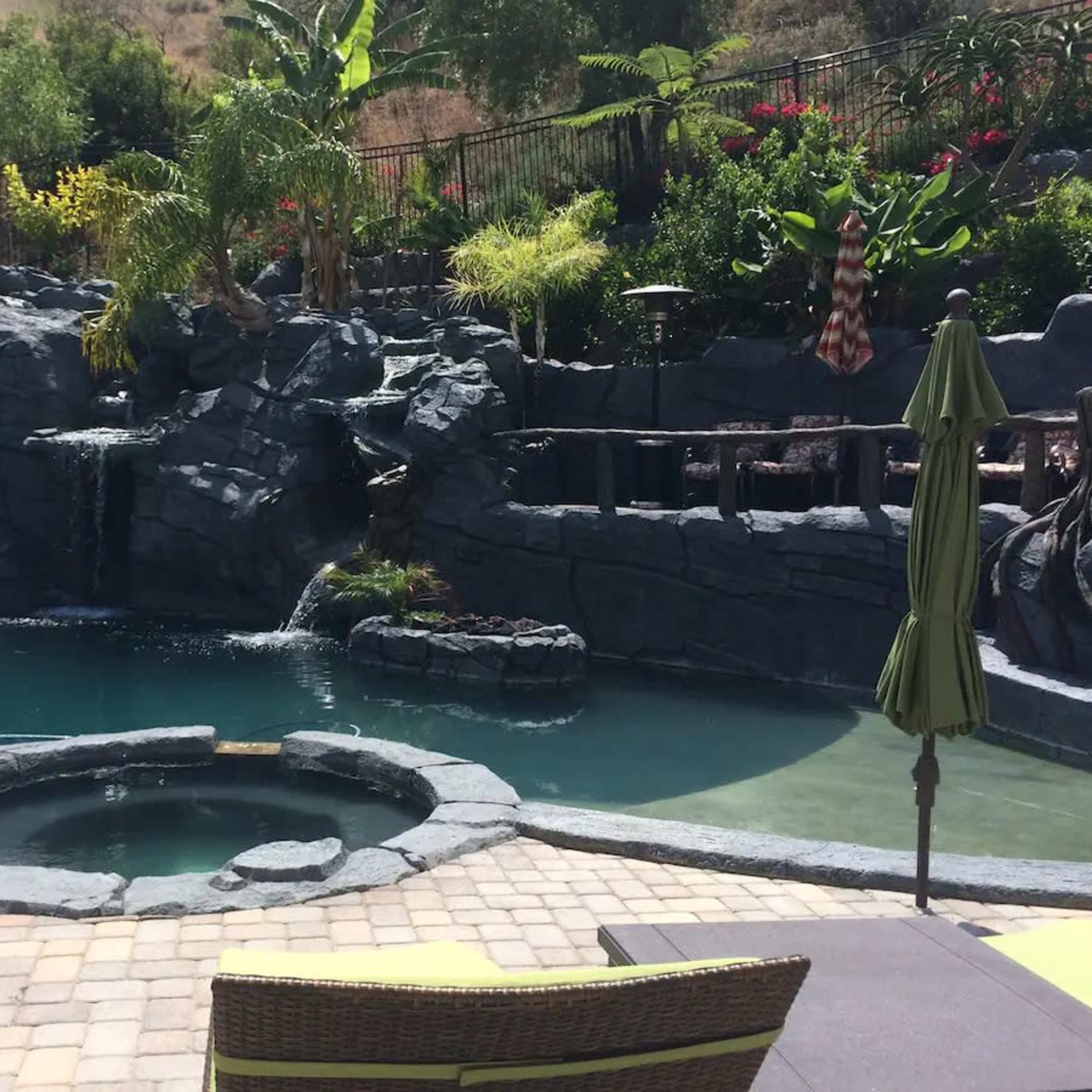 A landscaped pool area featuring a rock waterfall, a hot tub, and lush greenery.