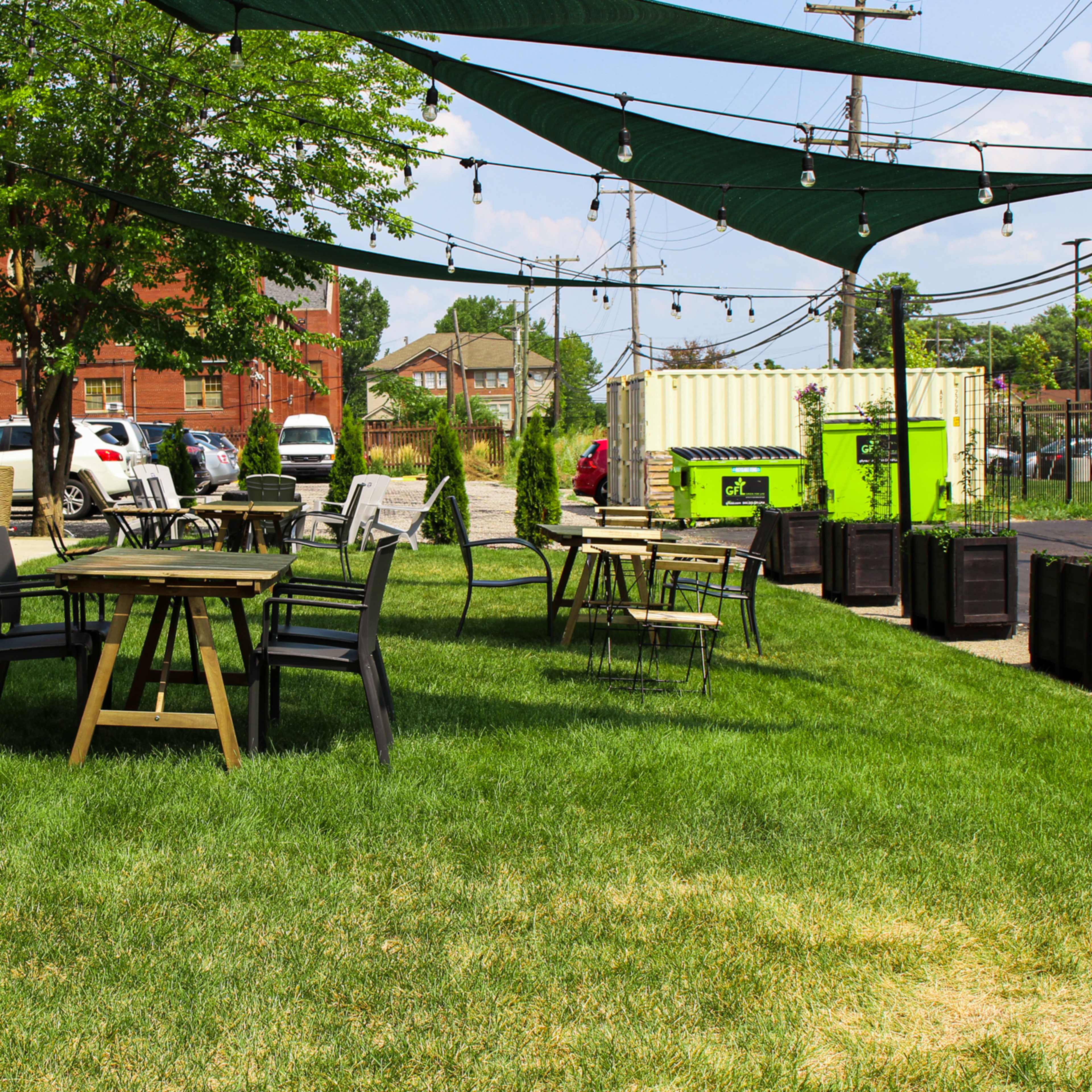 A grassy outdoor seating area with wooden tables and chairs, shaded by green canopies and surrounded by a brick building and parked vehicles.
